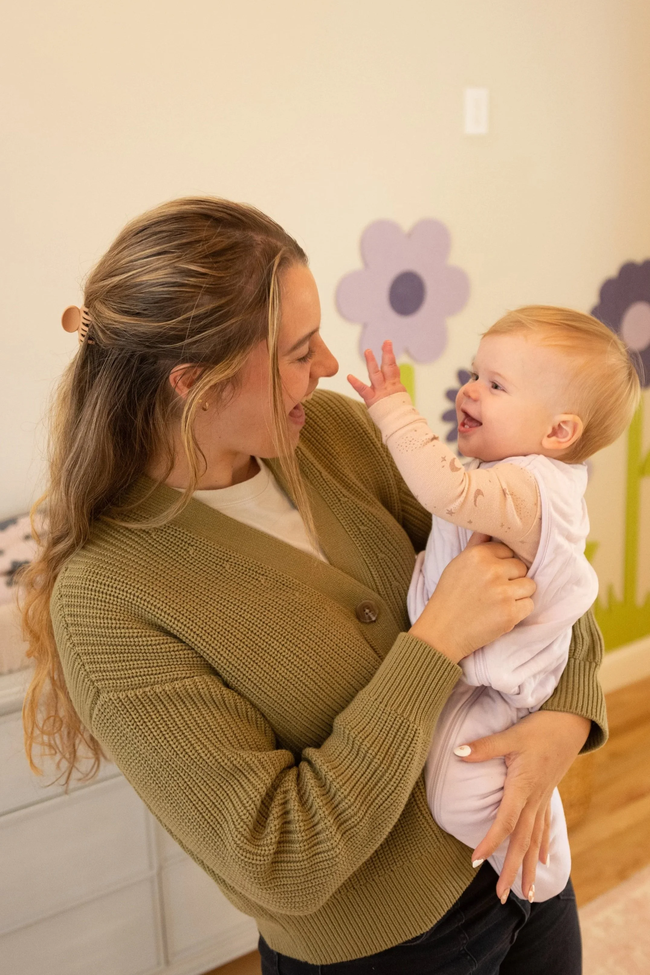 A woman holding a smiling toddler girl in a room decorated with purple flower wall decals.