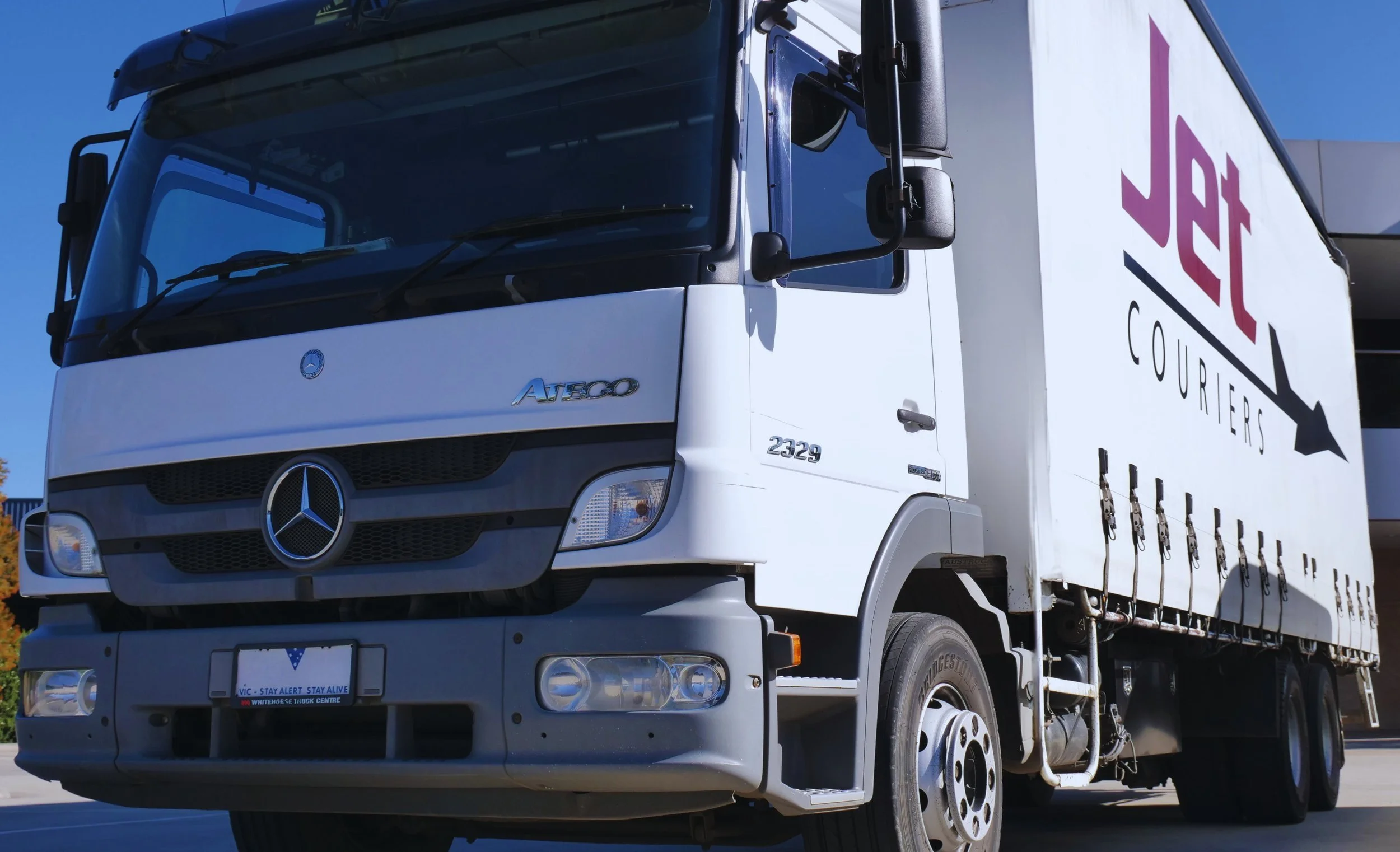 White courier truck with a Mercedes-Benz logo on the front, carrying JET Couriers branding on the side, parked outdoors under a clear blue sky.