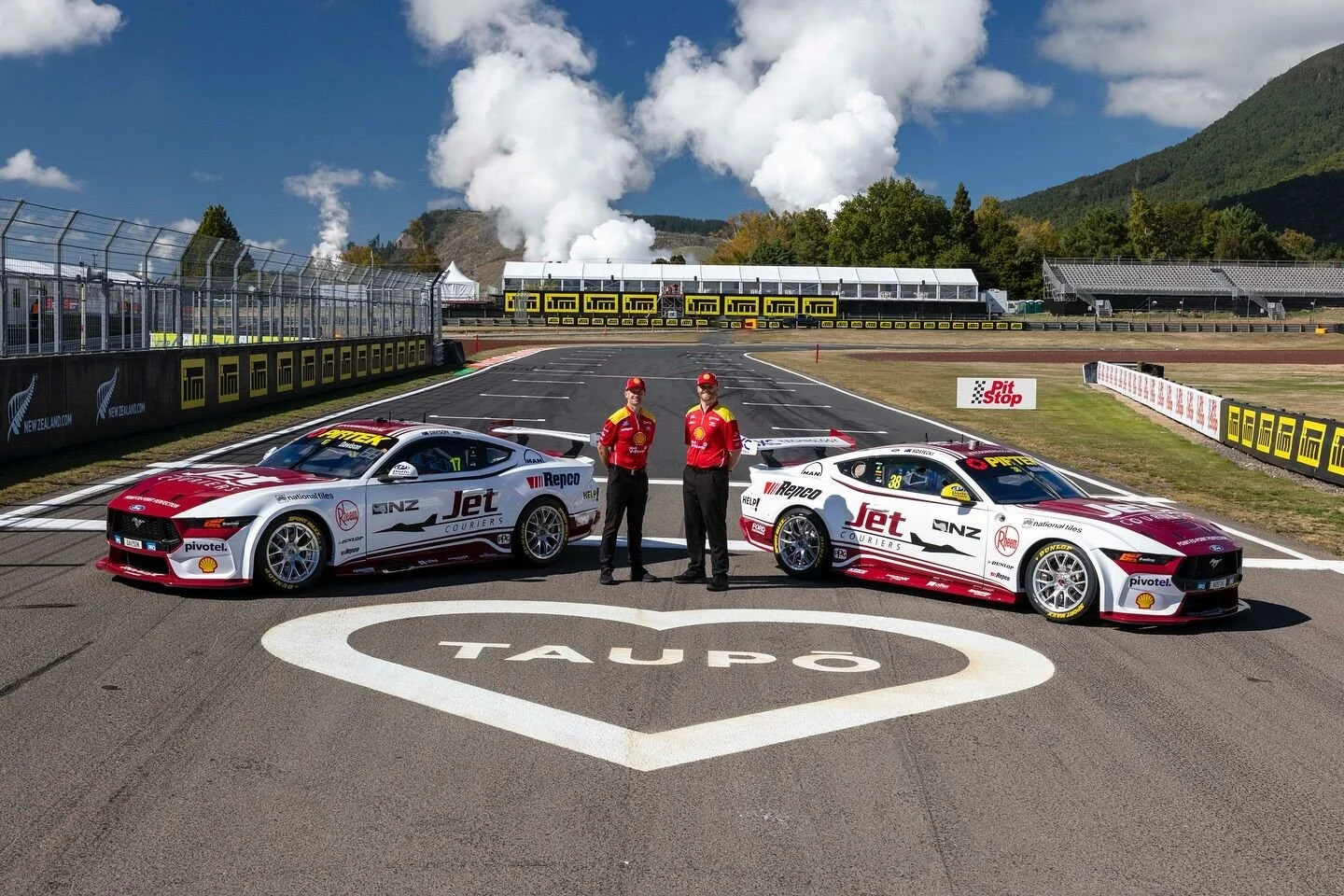 Two race cars and two crew members on a racetrack with mountains and racing infrastructure in the background.