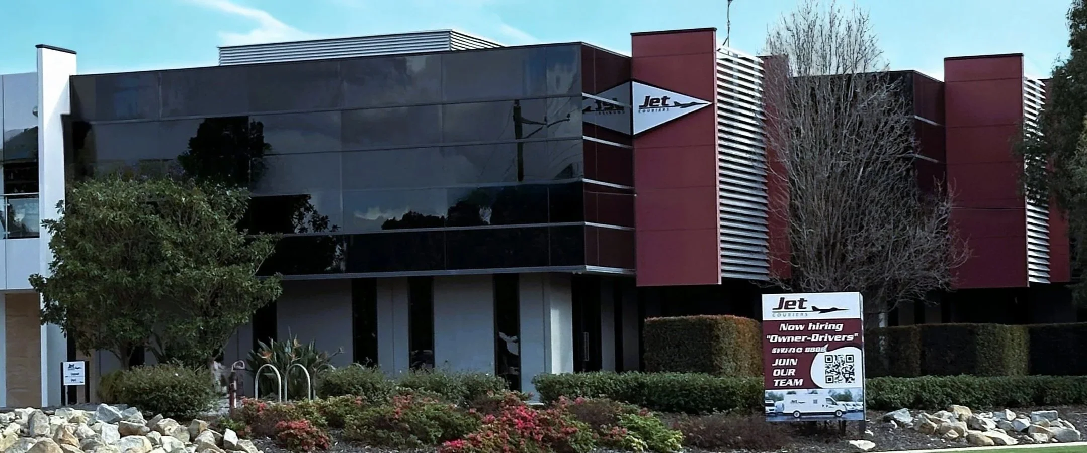 Modern building with reflective glass windows, red and white architectural accents, surrounded by landscaped bushes and trees, with a sign advertising job openings for a courier company.