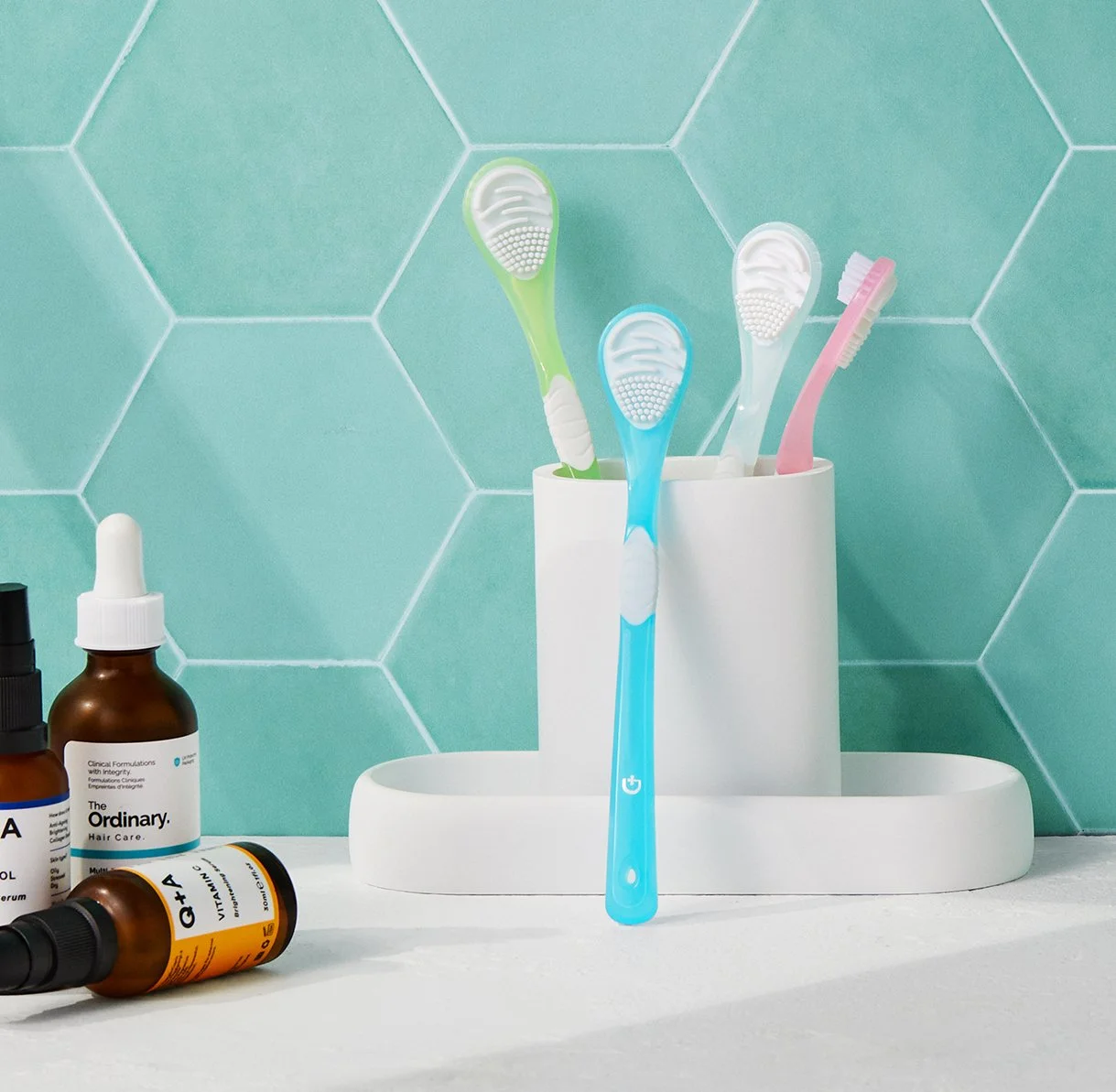 Four colorful toothbrushes in a white holder against a teal tiled wall, with skincare bottles on a white surface nearby.