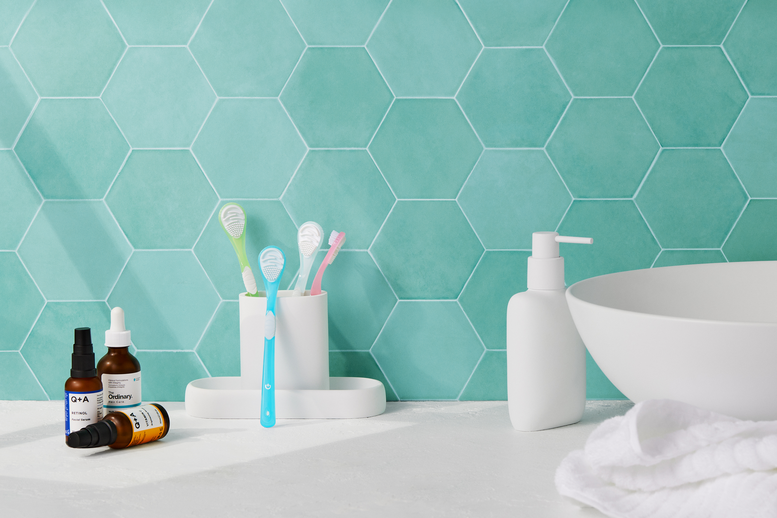 Bathroom countertop with toothbrushes in a holder, skincare bottles, a soap dispenser, a bowl, and a towel against a teal hexagonal tiled wall.