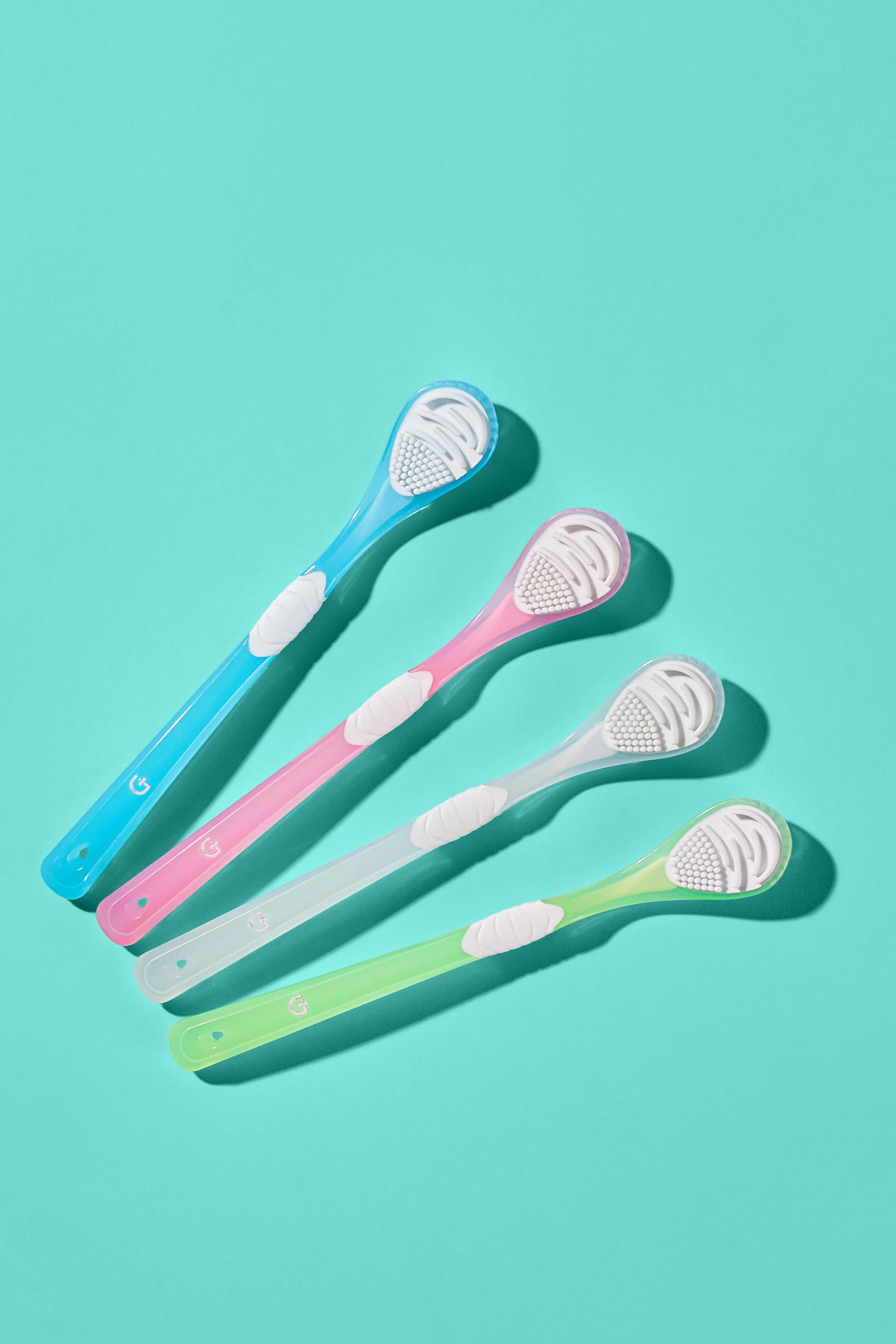 Four colorful toothbrushes in blue, pink, white, and green are arranged on a mint green background with shadows.