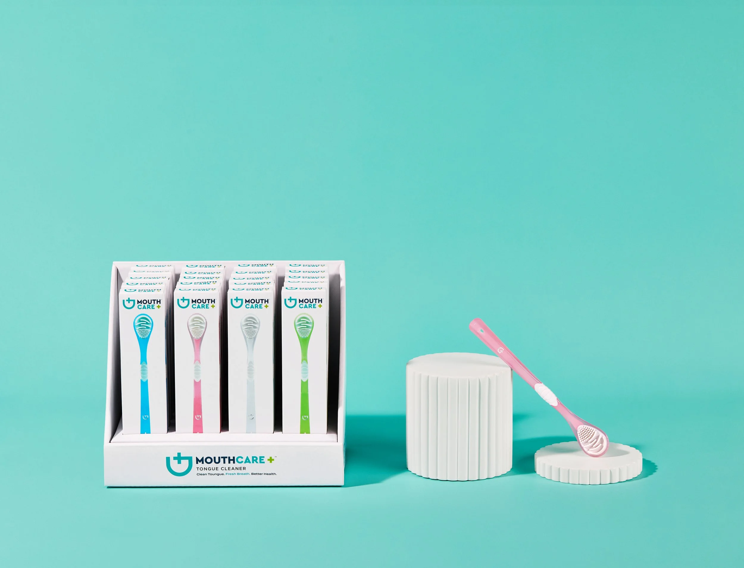 Display of four toothbrushes in a white box with a mint green, pink, white, and green design, a pink toothbrush laying on a white textured container, all against a mint green background.