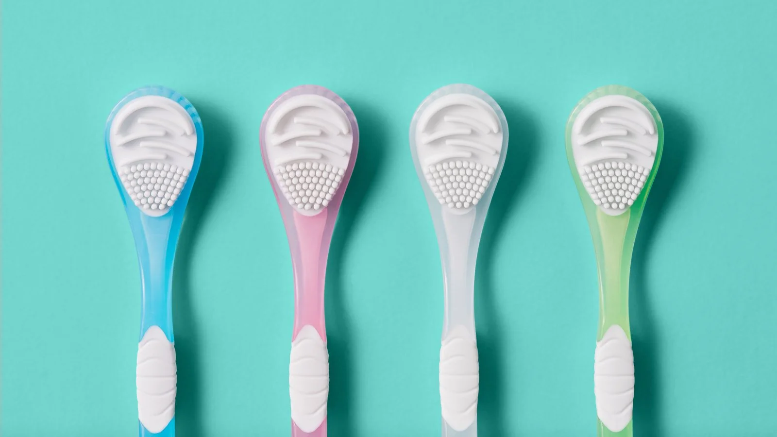 Four toothbrushes with blue, pink, white, and green handles arranged on a light turquoise background.