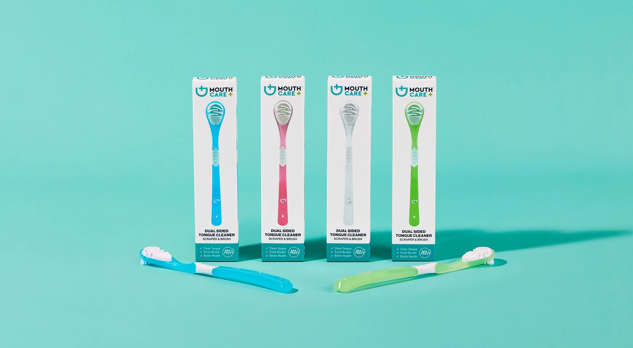Four boxed dual-sided tongue cleaners with matching toothbrushes laid out in front, each in different colors: blue, pink, white, and green, on a teal background.