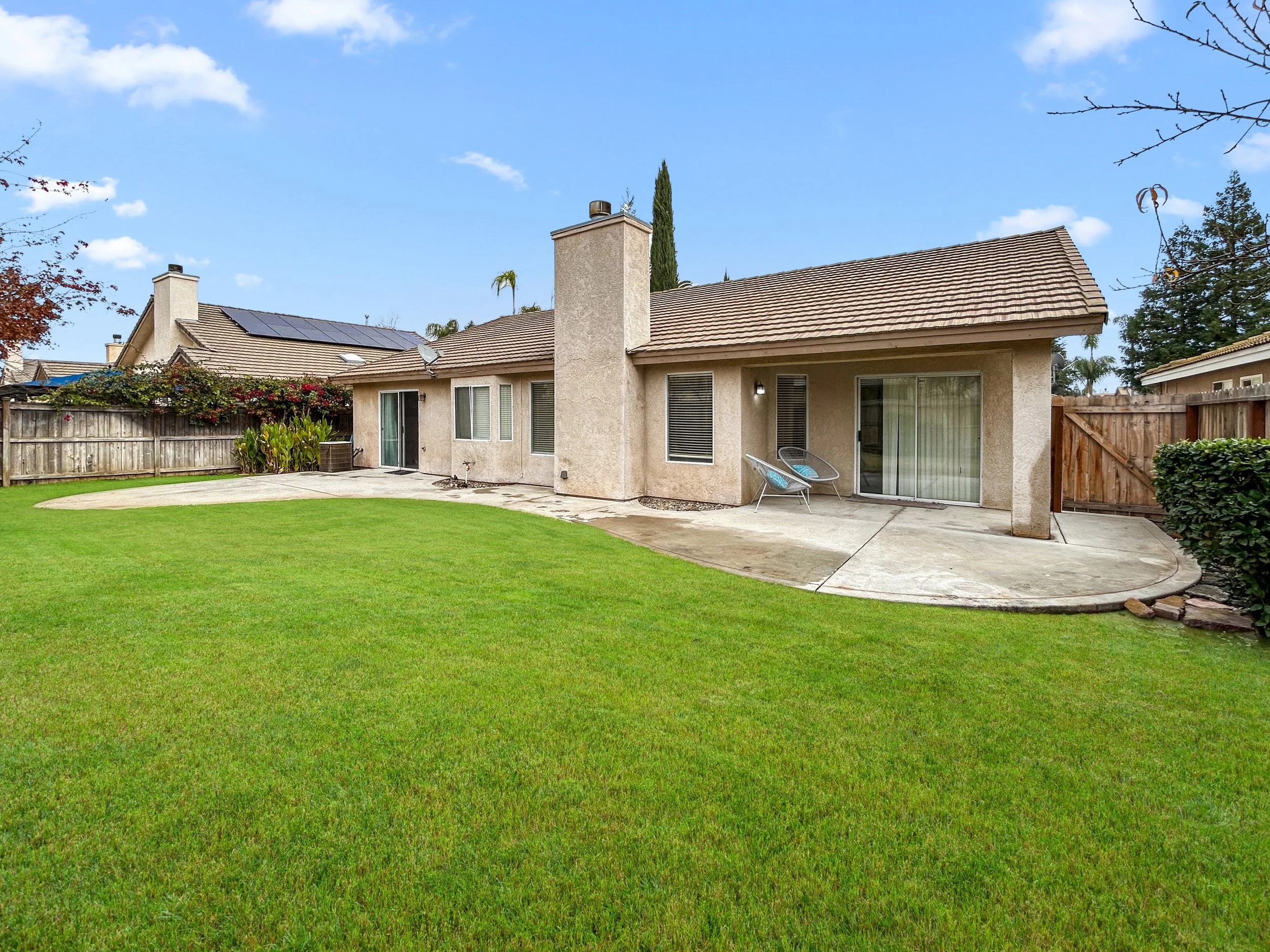 Backyard view of a beige stucco house with a tiled roof, sliding glass doors, and a patio area, surrounded by a green lawn and a wooden fence, with neighboring houses and a blue sky in the background.