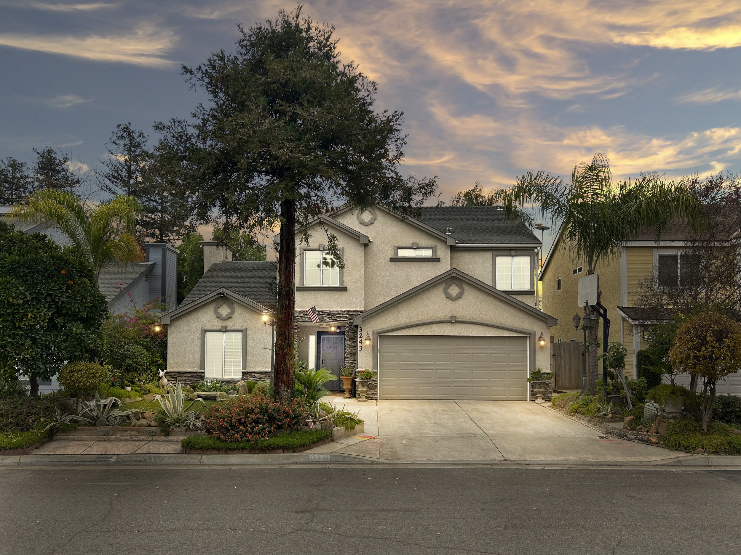 A two-story suburban house with a gray garage door, surrounded by landscaped garden, trees, and neighboring homes at dusk under a partly cloudy sky.