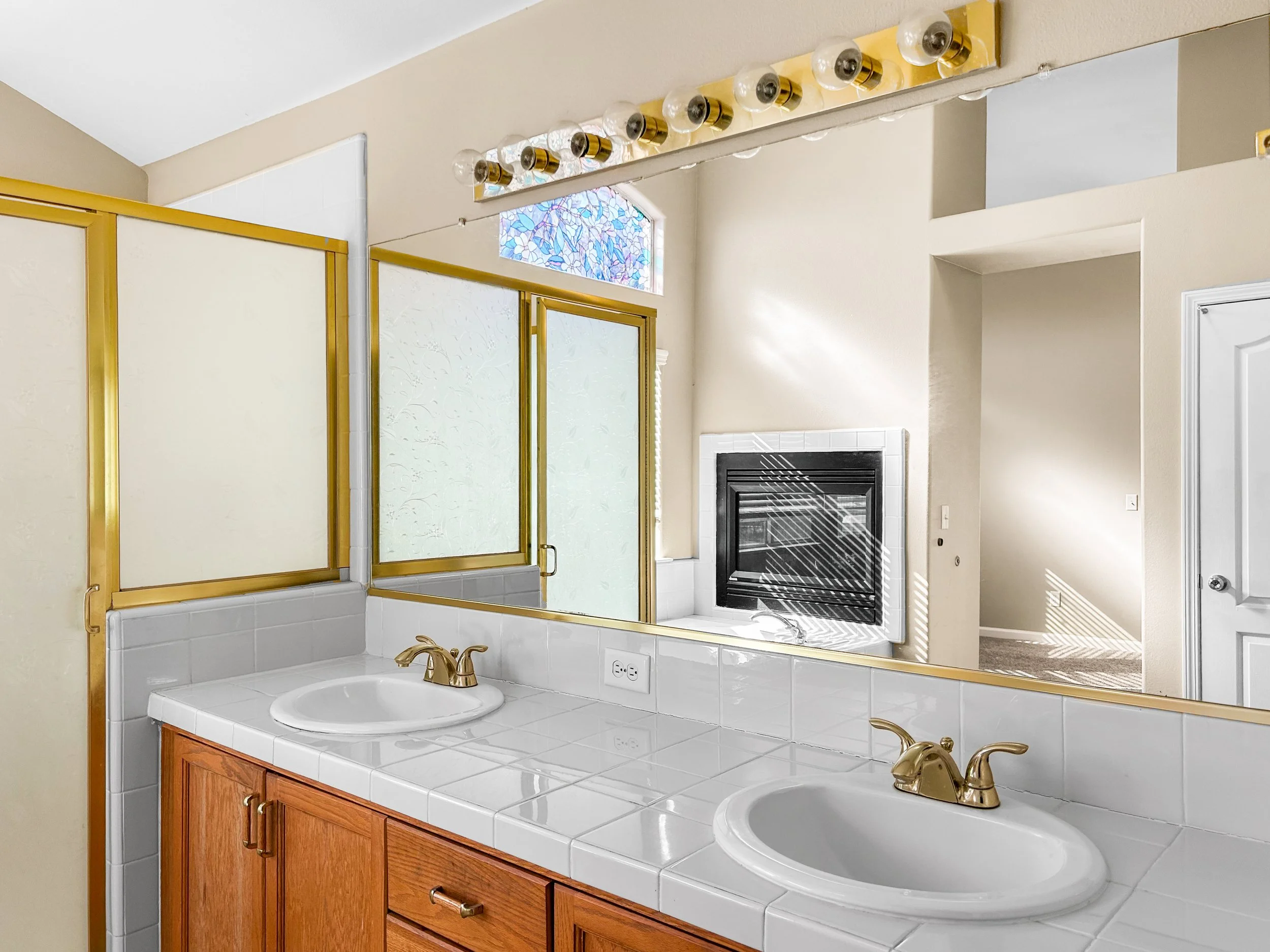 Bathroom with dual sinks, gold fixtures, white tiled countertop, large mirror, shower with glass door, fireplace, and a small stained glass window.