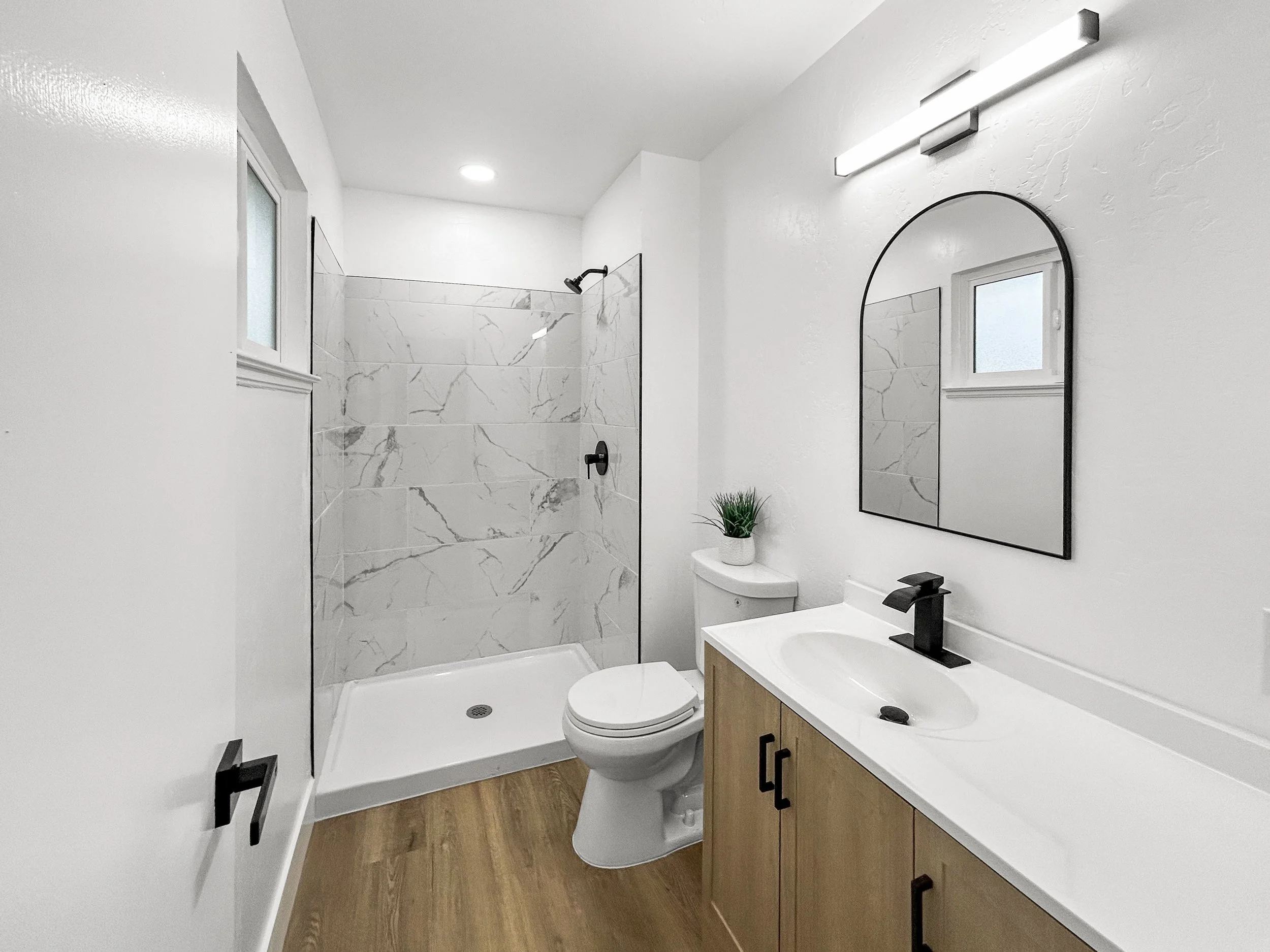 Modern bathroom with walk-in shower, white walls, wooden vanity with black hardware, oval mirror, rectangular light fixture, small window, black faucet, green plant, toilet, wood-look flooring.
