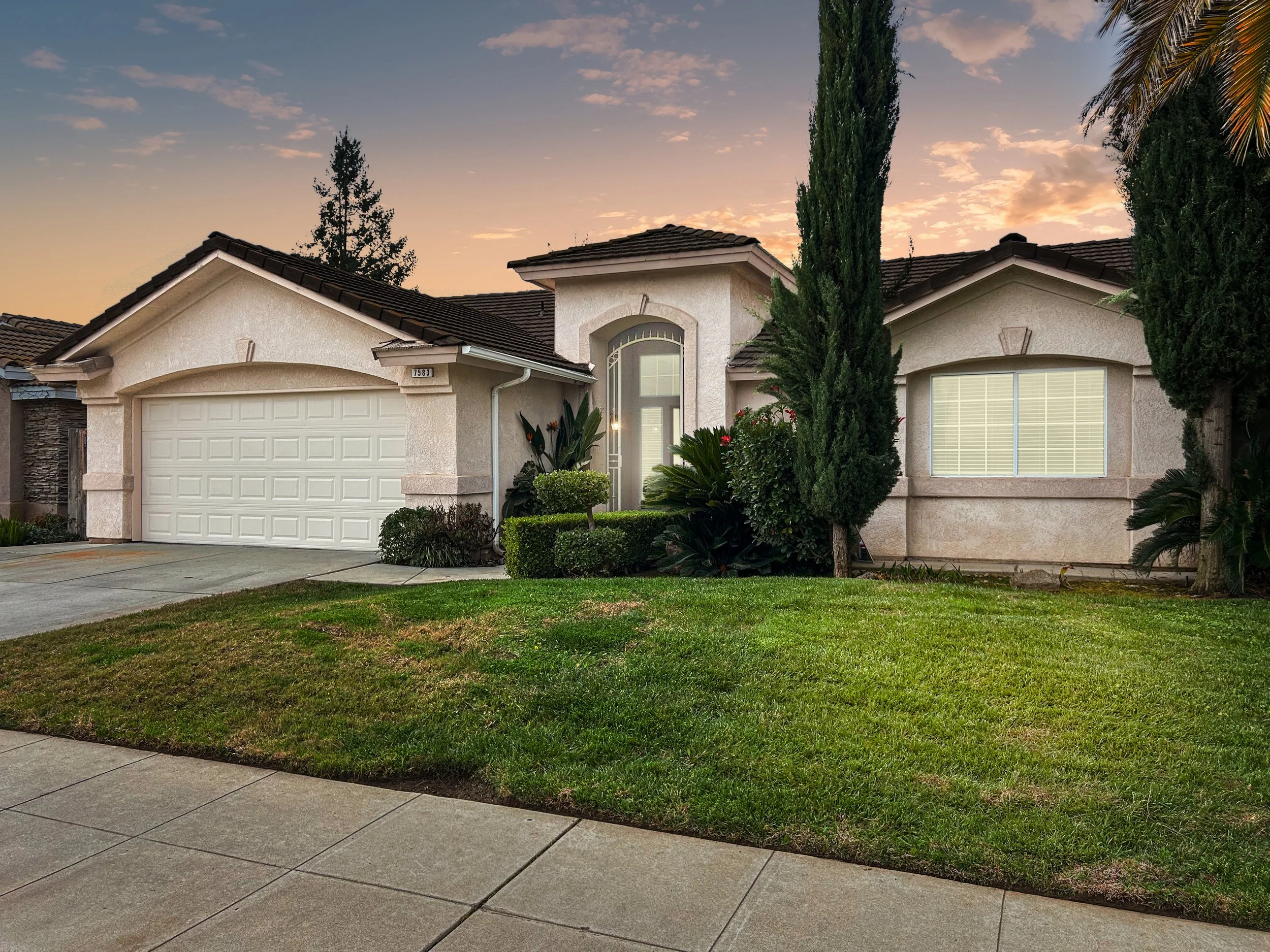A single-story suburban house with a white garage door, beige exterior walls, and a brown roof, with a well-maintained front lawn, trees, and shrubs during sunset.