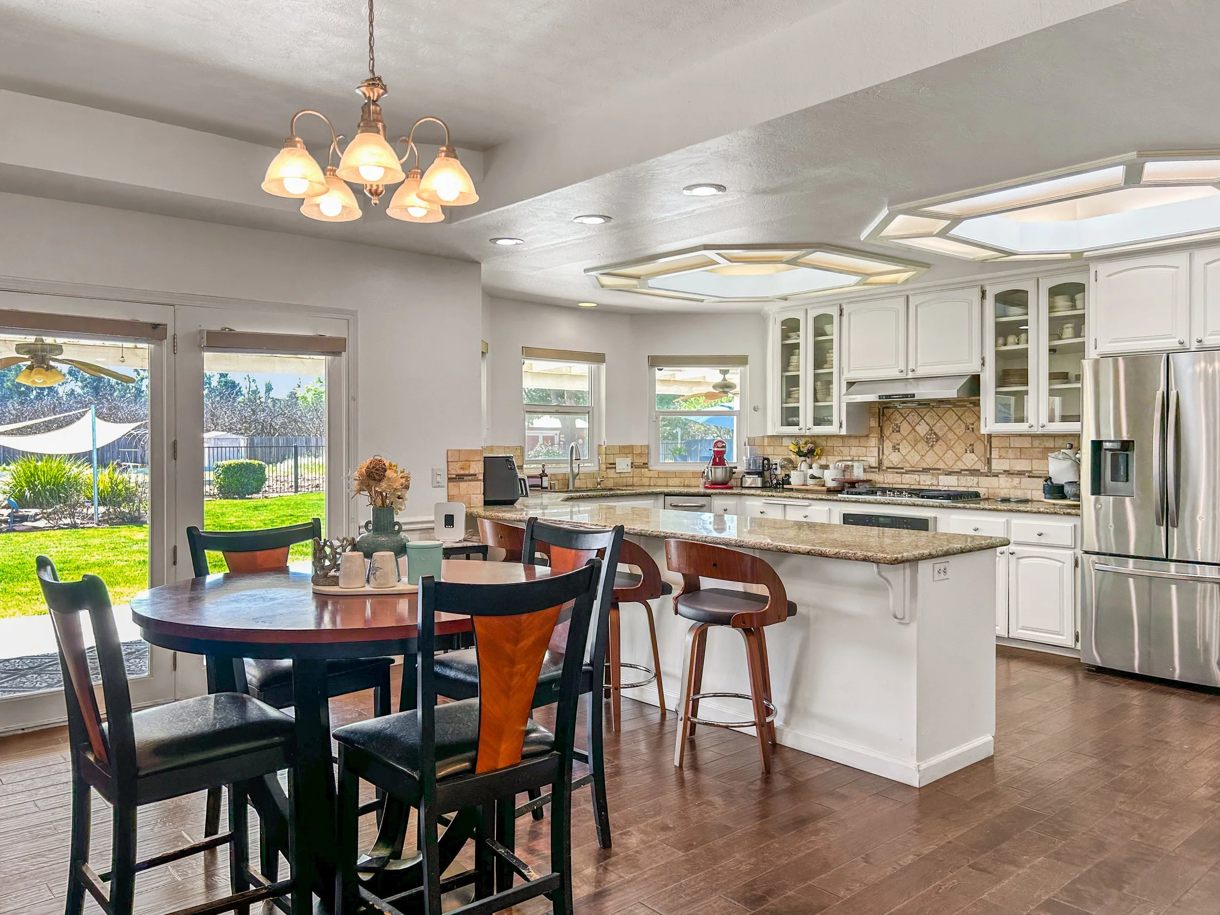 Bright kitchen with white cabinets, granite countertops, stainless steel refrigerator, and a round dining table with six chairs near sliding glass doors leading to a backyard with grass and bushes.