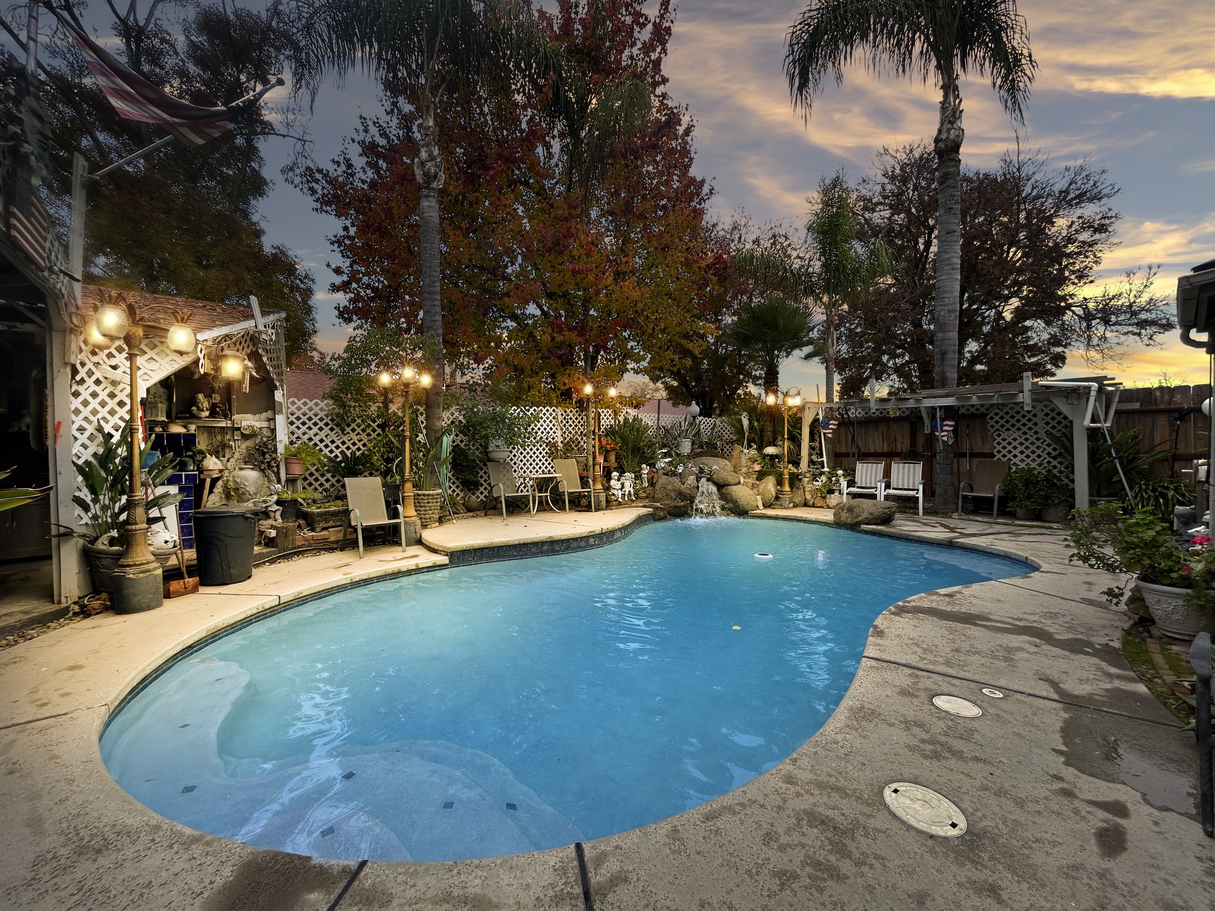 A backyard swimming pool at sunset with trees, string lights, and outdoor seating.