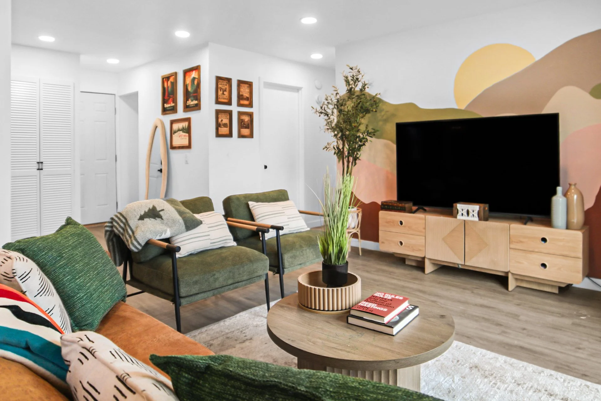 A modern living room with a colorful mural on the wall, a wooden TV stand with a large flat-screen TV, a natural wood coffee table, and multiple green and orange upholstered chairs and couches decorated with patterned pillows and blankets. Decorative