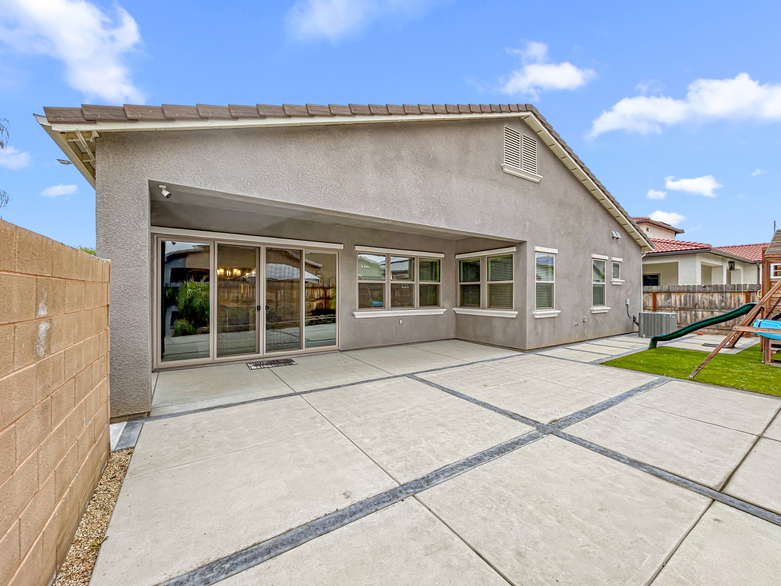 Backyard patio with concrete slabs, sliding glass door, windows, a grassy area with a slide, and a fenced backyard under a partly cloudy sky.