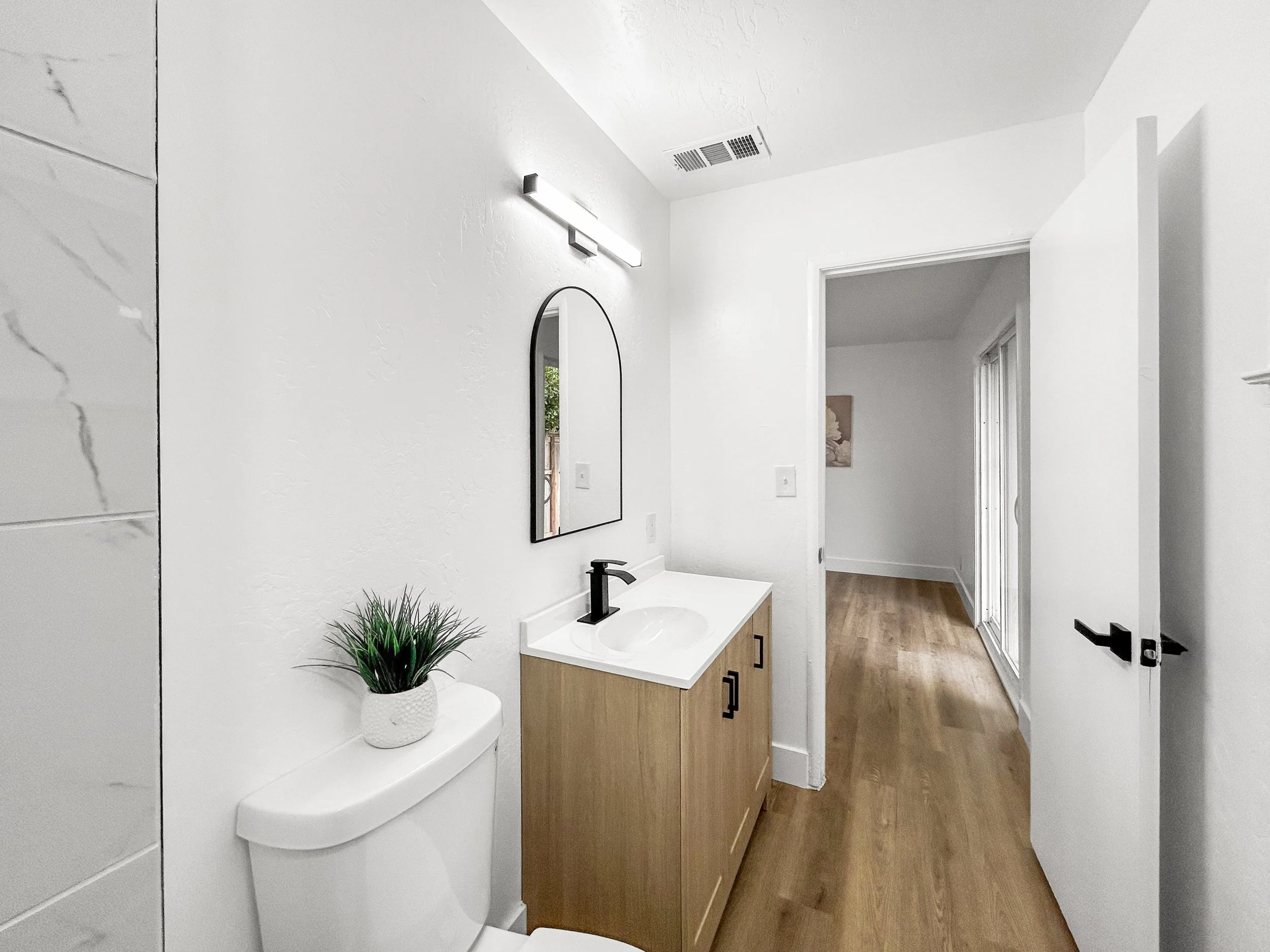 Modern bathroom with white walls and wooden floor. Includes a toilet with a small potted plant on top, a vanity with a black faucet, a mirror, and a door leading to another room.