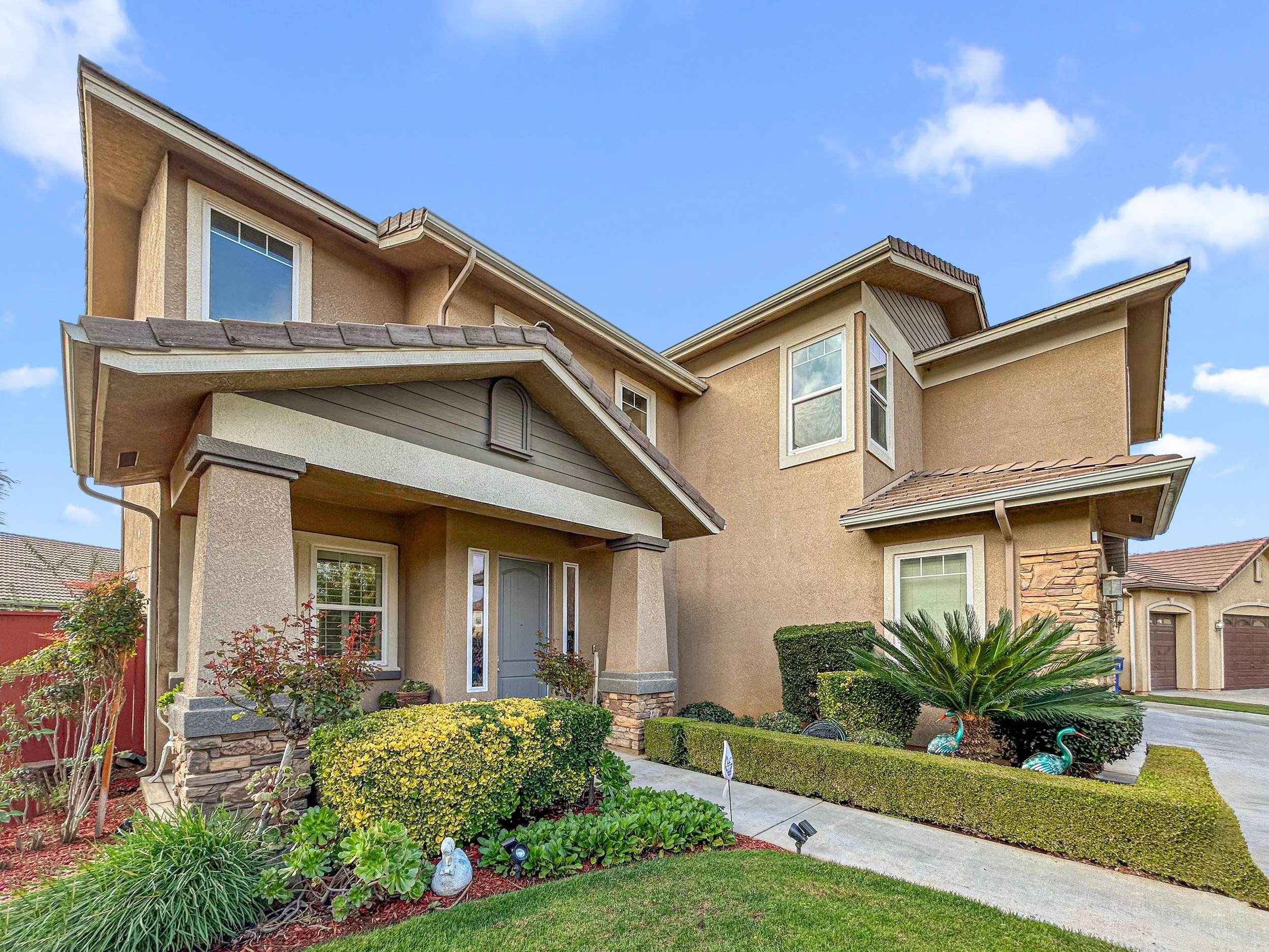 A two-story beige house with a landscaped front yard featuring shrubs, a small palm tree, and decorative garden ornaments, under a blue sky with some clouds.