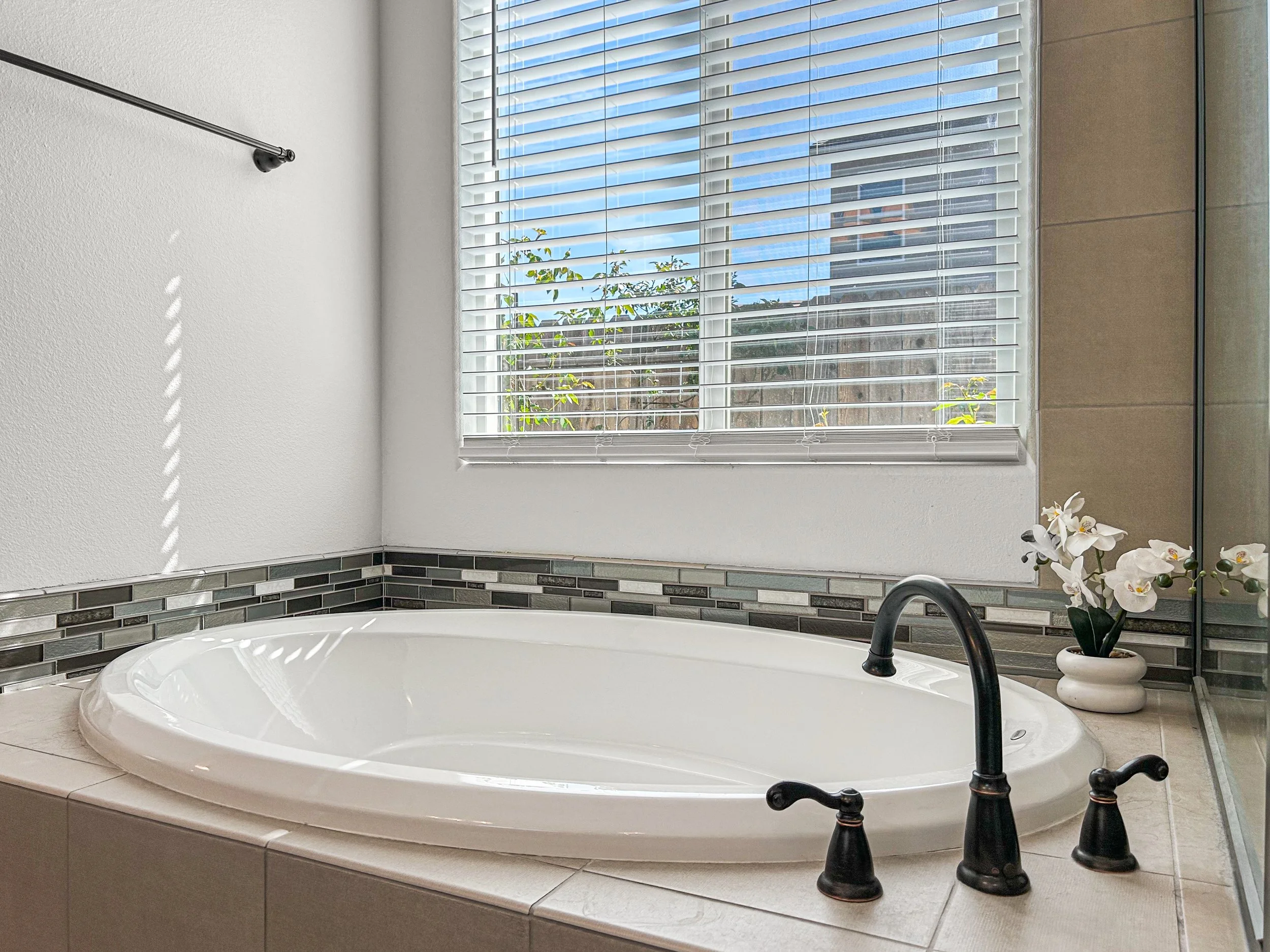 A bathroom with a large bathtub, black faucet, decorative tile border, a window with blinds, and a potted orchid plant.