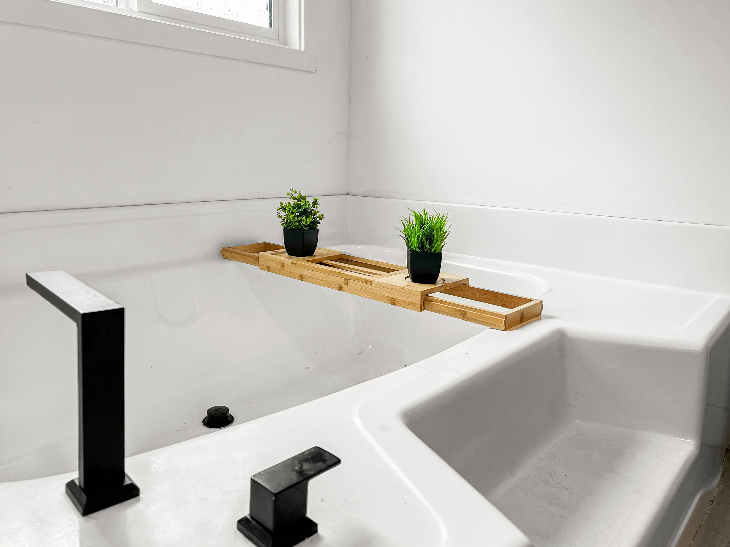 A white bathtub in a bathroom with a wooden tray holding two black pots with green plants, and a black faucet.