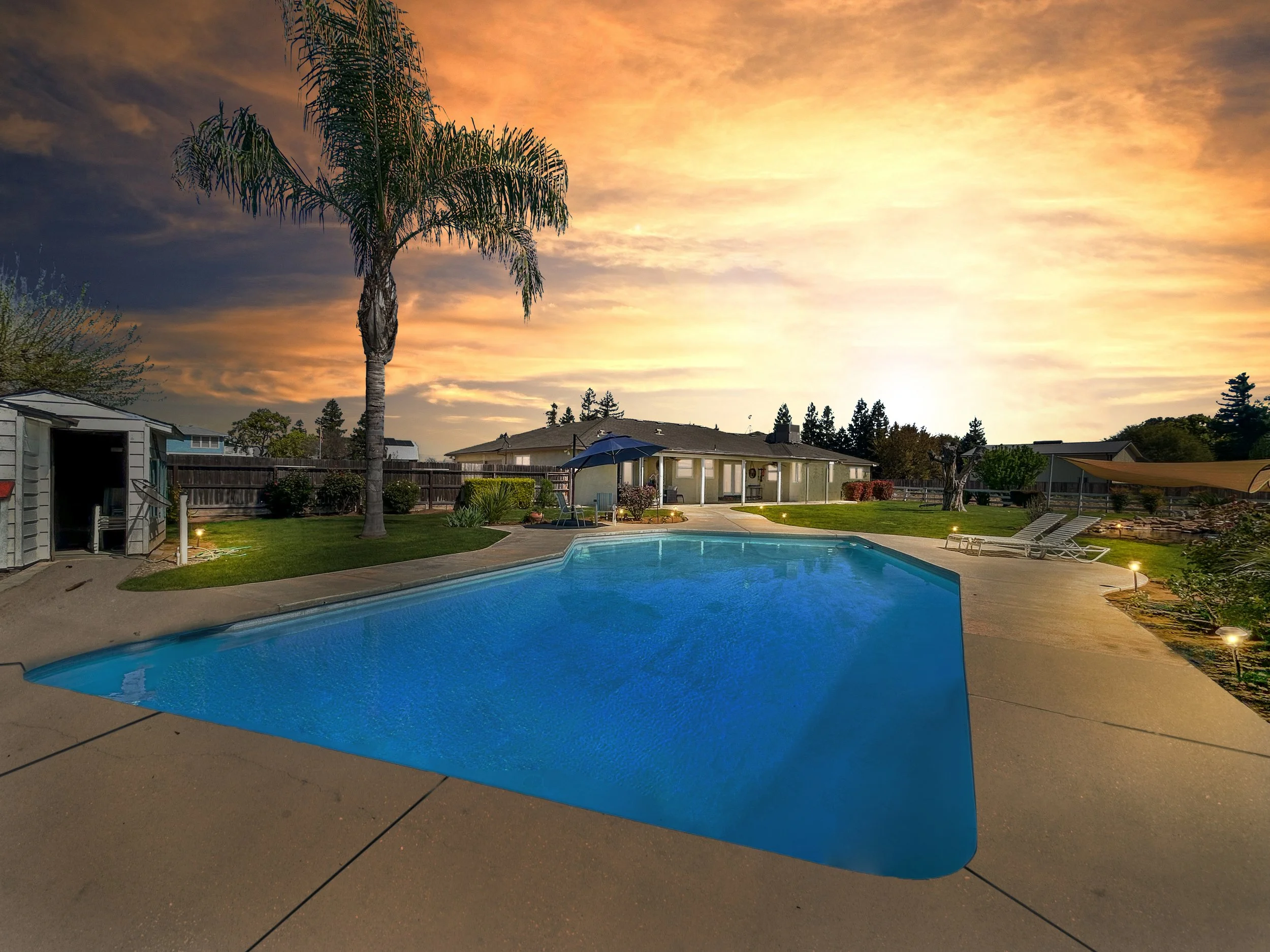 A backyard swimming pool at sunset with lounge chairs, a palm tree, a shed, and a house with a covered patio. The sky is orange and pink with some clouds.