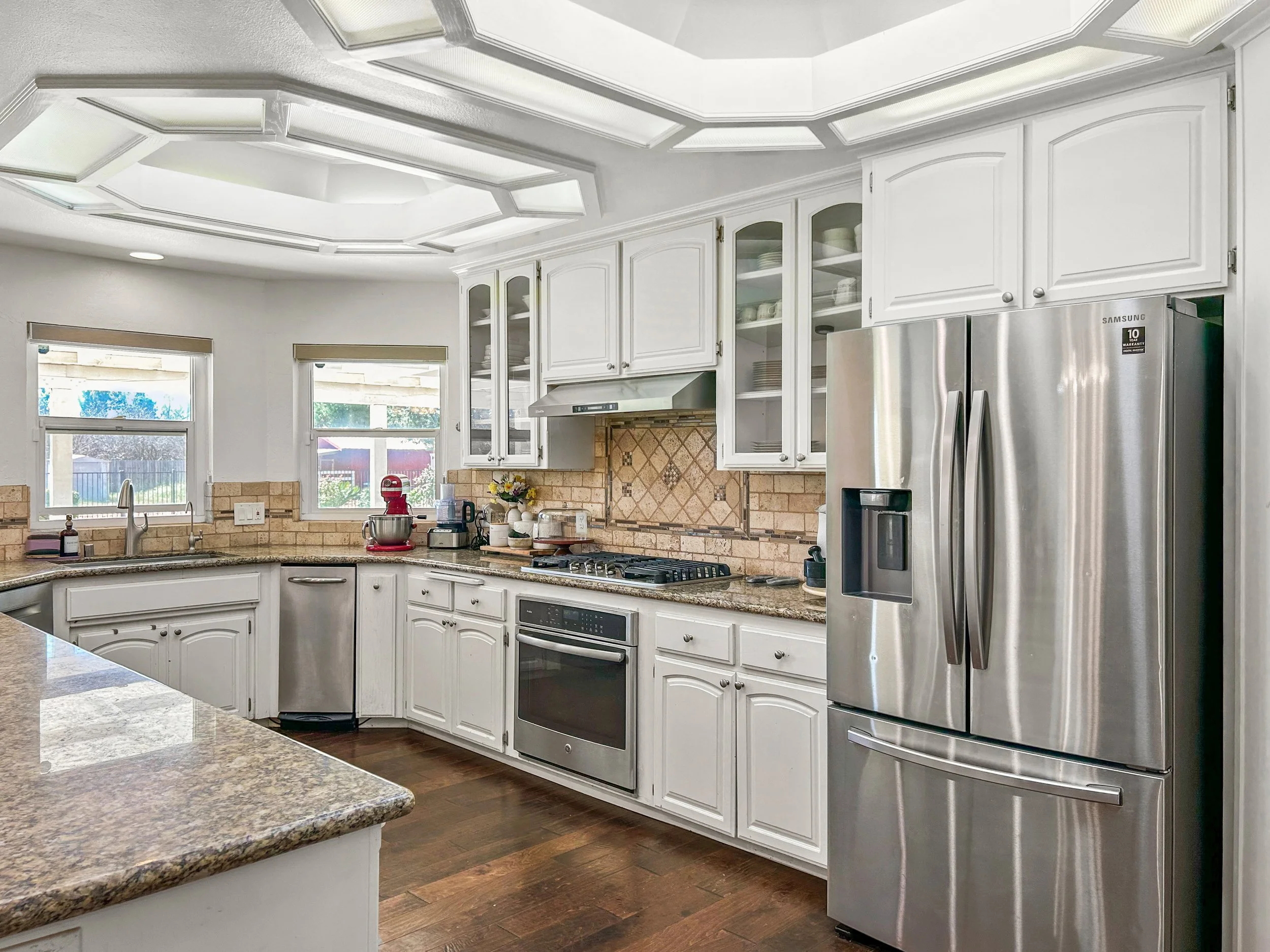 Modern kitchen with white cabinets, granite countertops, a stainless steel refrigerator, oven, and microwave, and windows letting in natural light.