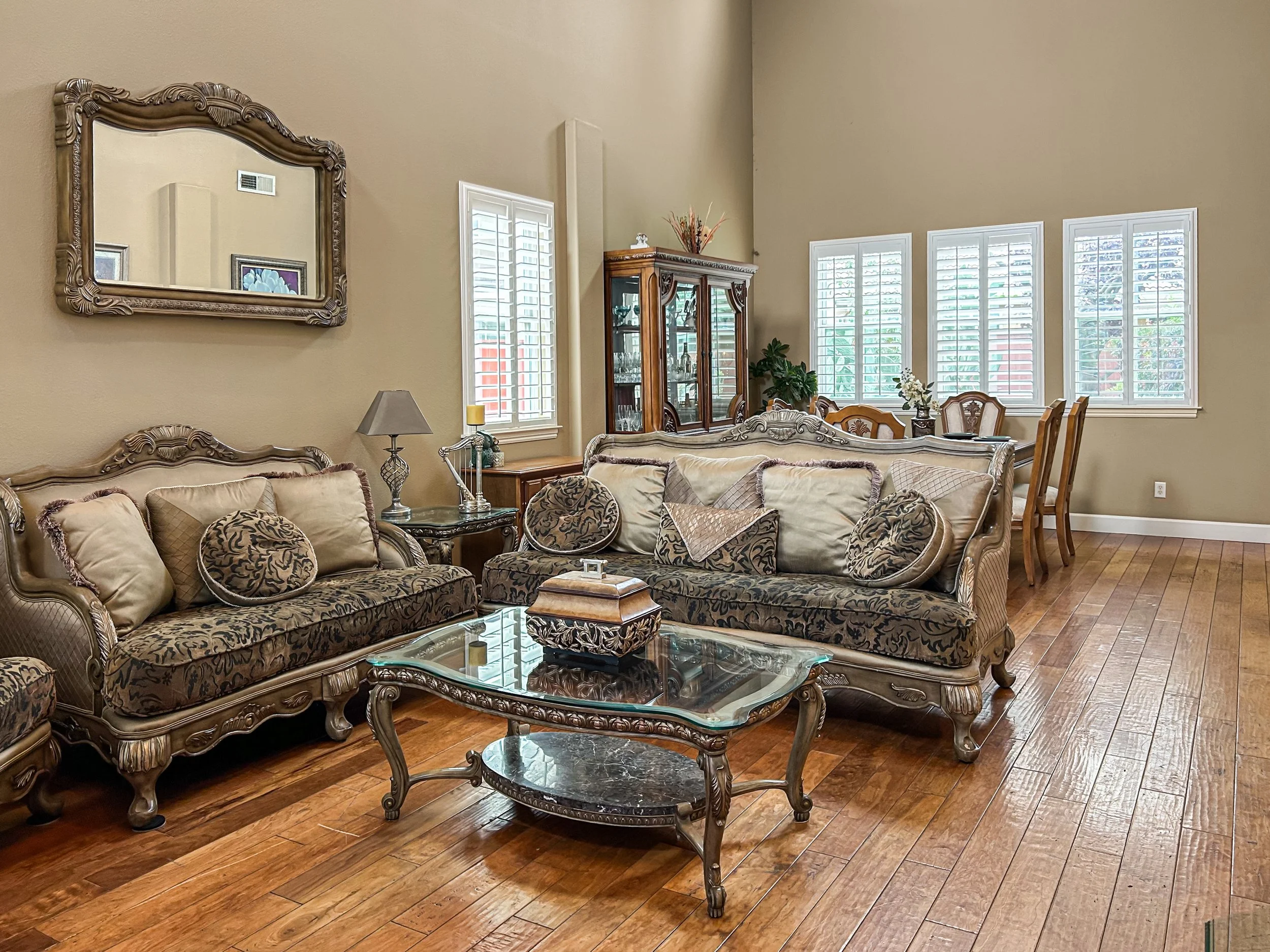 Living room with ornate beige and brown furniture, a glass coffee table, and hardwood floors. Multiple windows with white shutters and a dining area in the background.