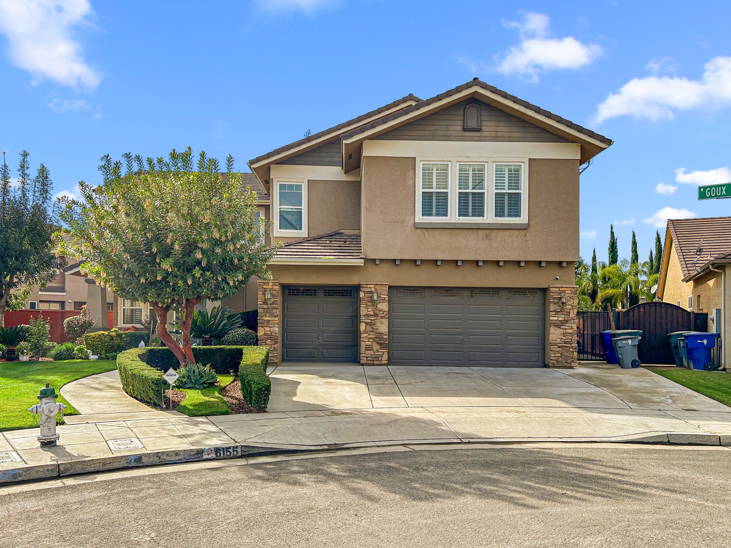 Two-story suburban house with three garage doors, stone accents, and trees in the front yard. Clear blue sky with a few clouds.