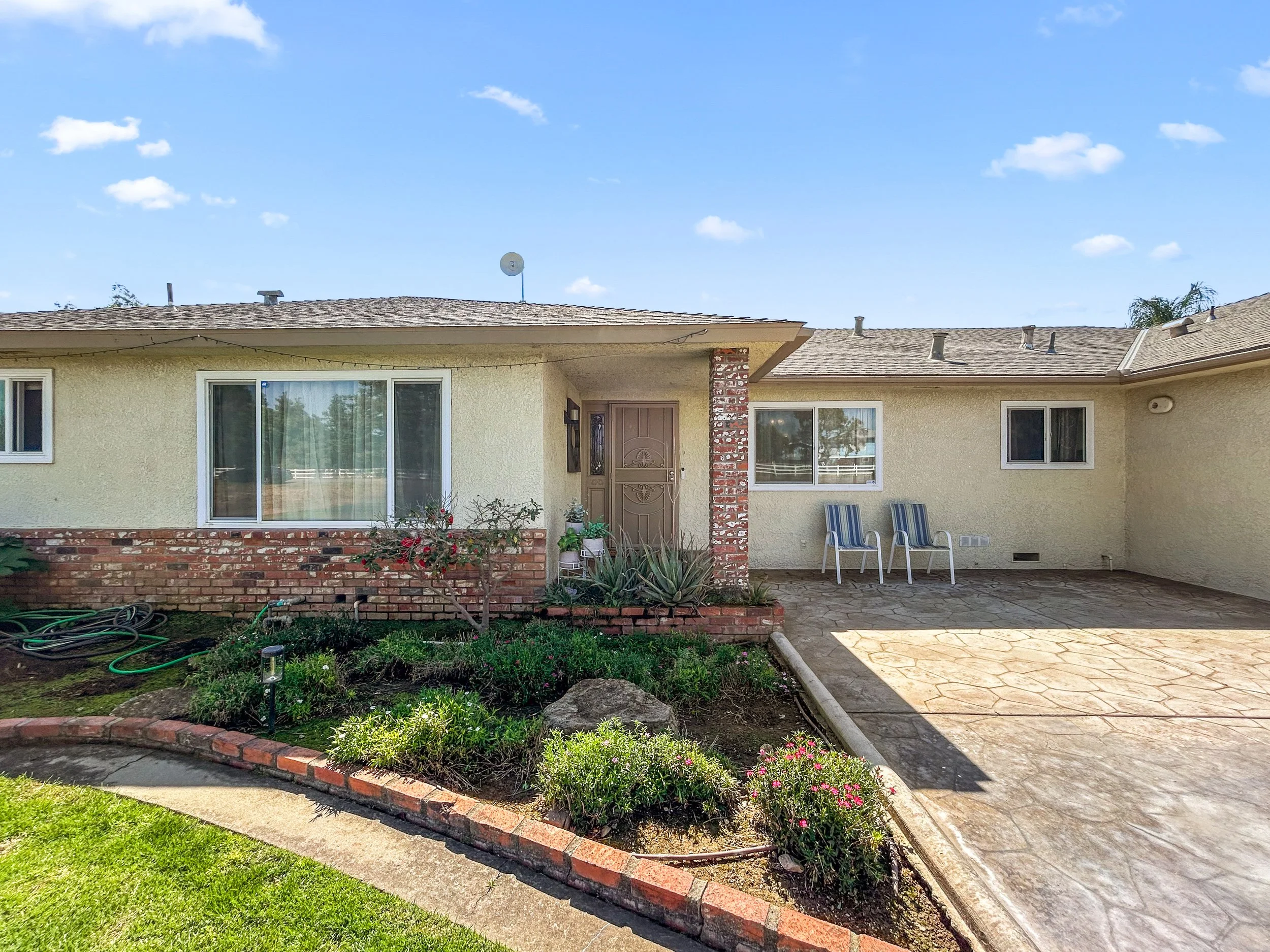 Exterior view of a single-story house with a brick border flower bed, two blue and white striped chairs on a concrete patio, large windows, and a brown front door. Clear sky with a few clouds.