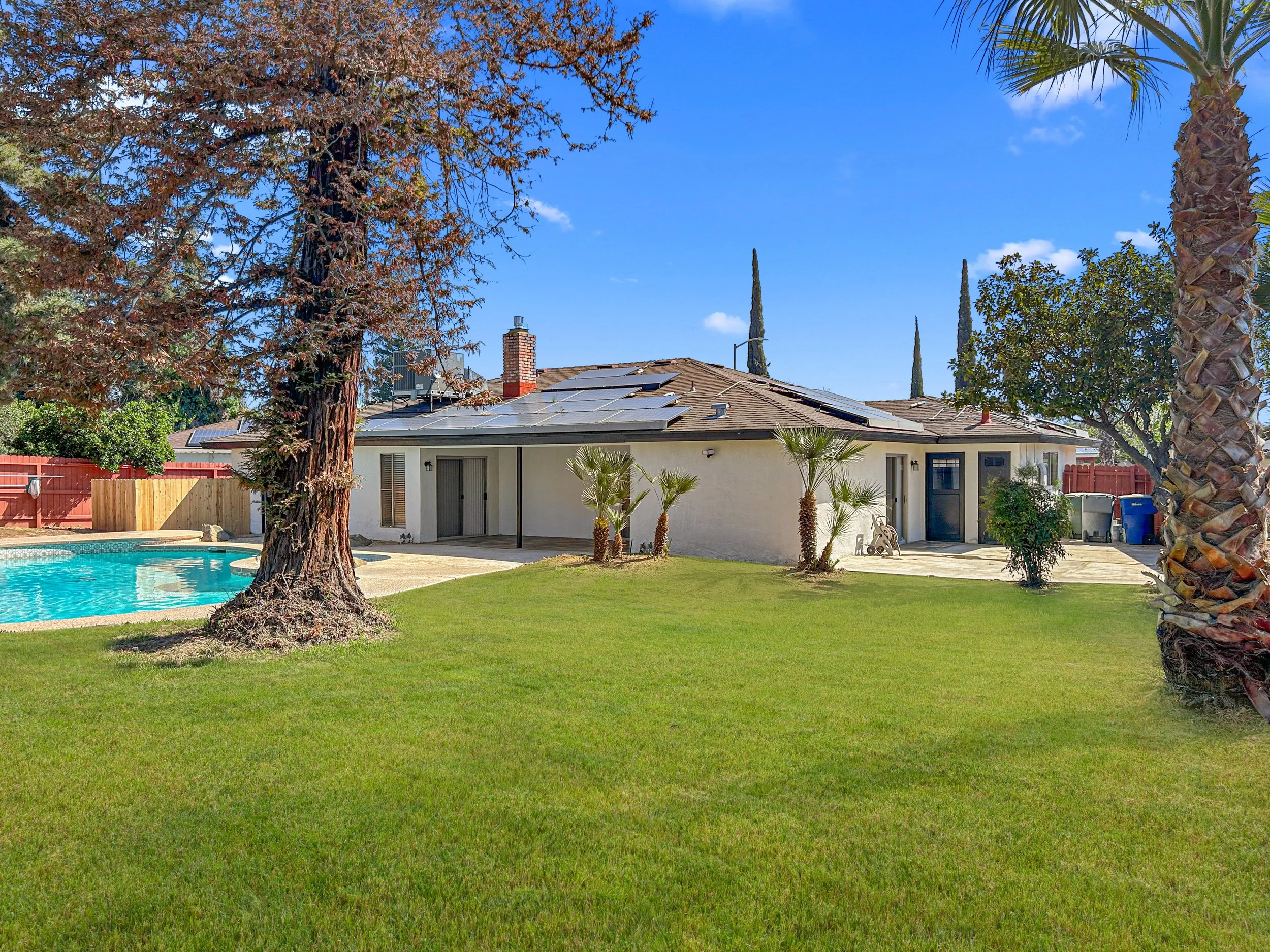 Backyard view of a house with a swimming pool, large trees, palm trees, and a well-maintained lawn on a sunny day.
