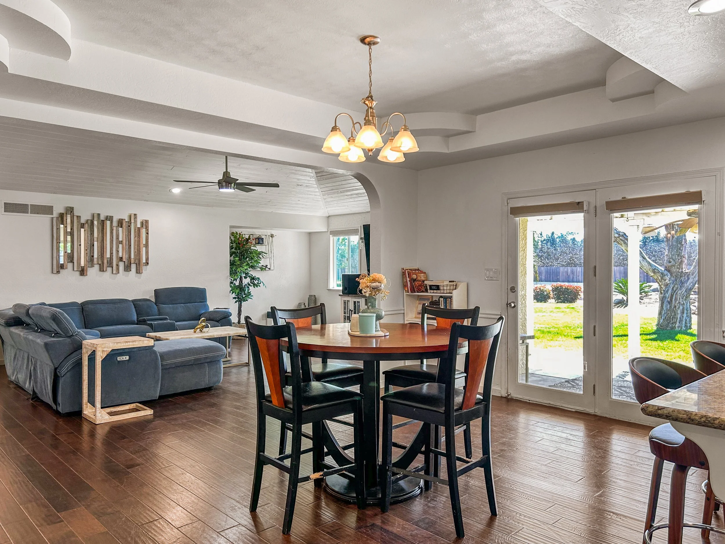 Living and dining area with wooden floor, blue sectional sofa, round dining table with black and brown chairs, glass double doors leading outside, ceiling fan, hanging chandelier, window with blinds, decorative artwork, indoor plants, and outdoor yar