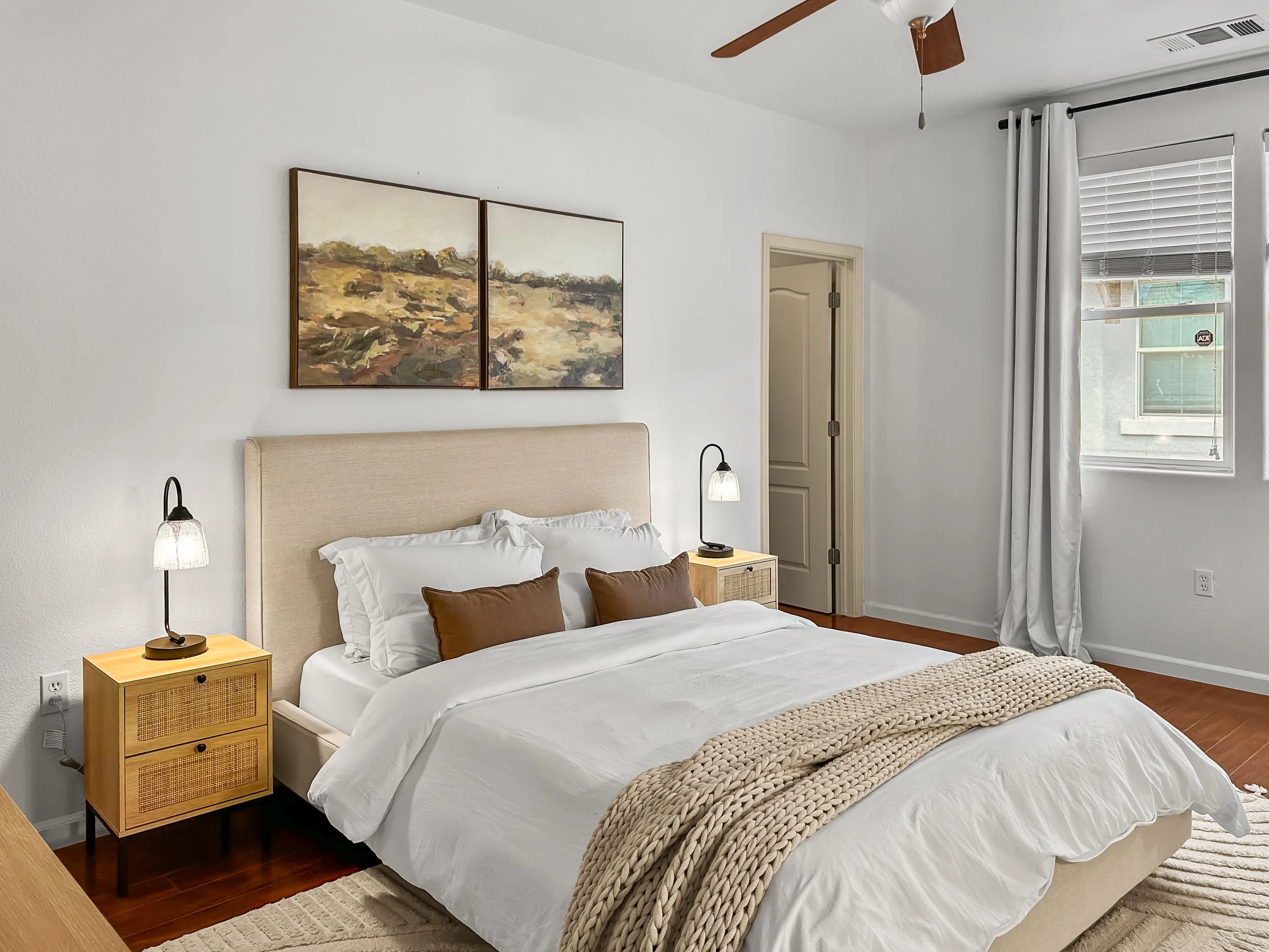 A bedroom with a bed, two wooden nightstands with lamps, white curtains, and a window. There is artwork on the wall above the bed and a ceiling fan.