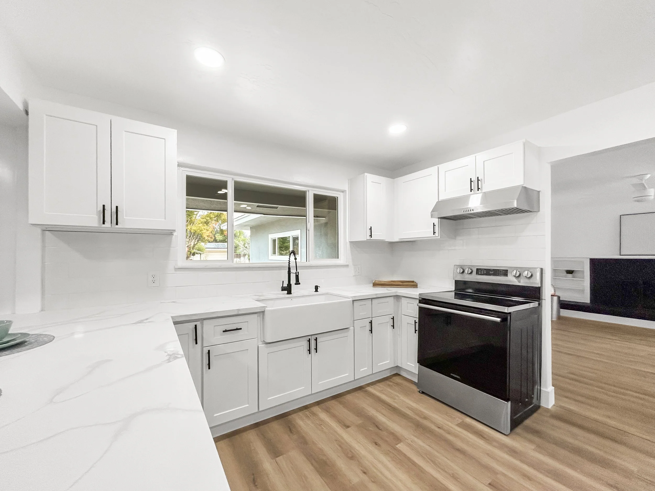 Modern kitchen with white cabinets, black hardware, stainless steel oven, black stove, white marble countertops, black faucet, window above sink, and wood flooring.