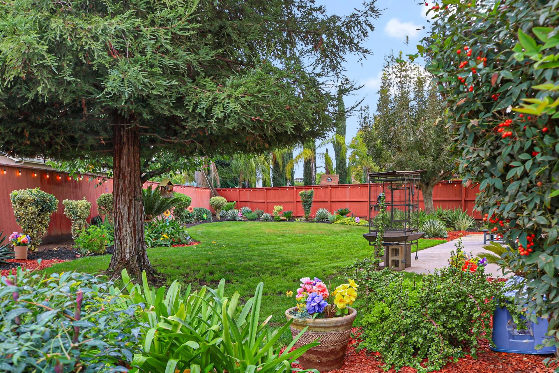A backyard garden with a large tree, well-manicured grass, colorful flowers, shrubs, a red wooden fence, a pathway, and garden decorations.