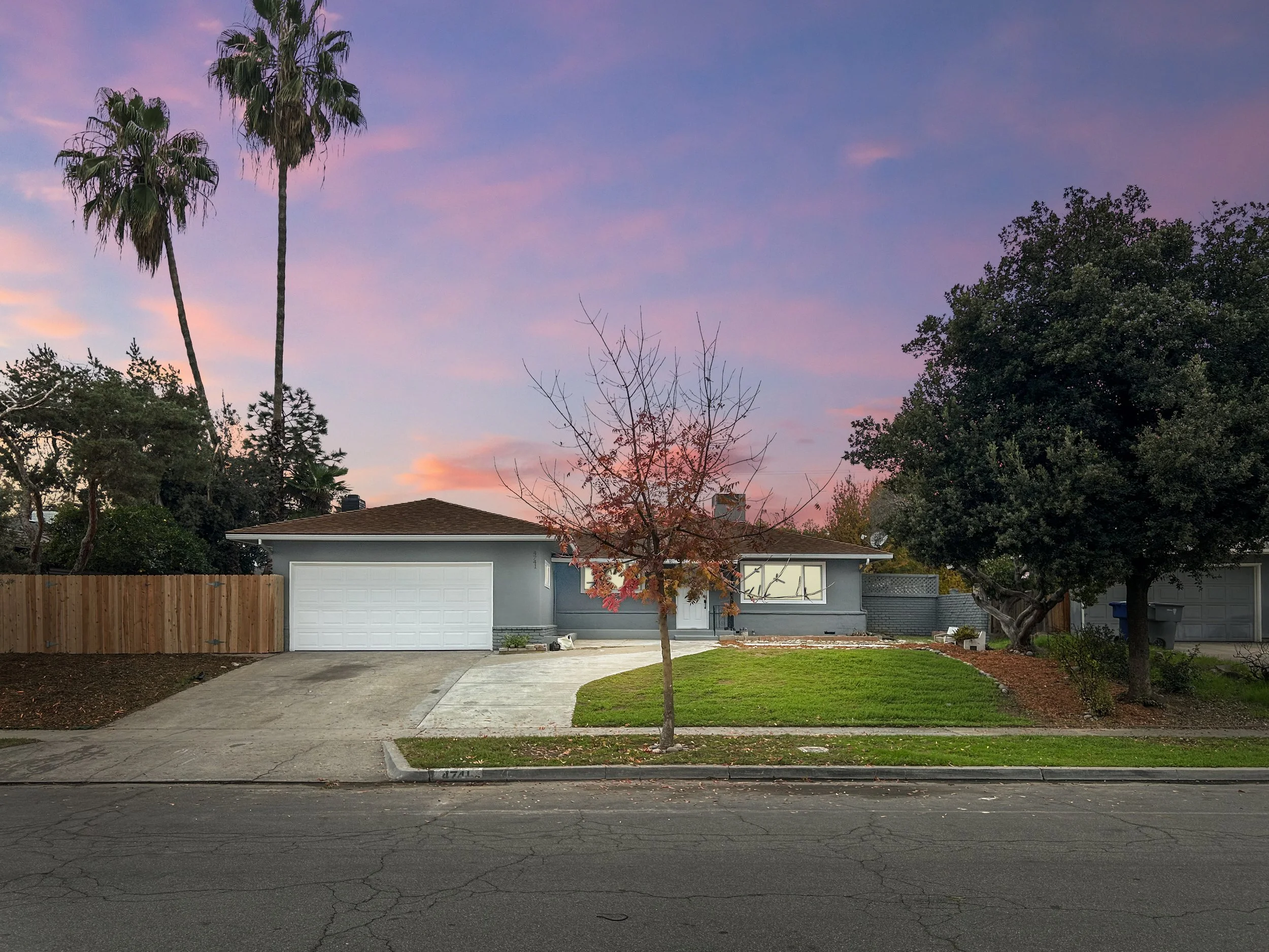 A suburban house with a light blue exterior, a brown roof, and a white garage door. There is a small tree with sparse leaves in front and a large tree on the right. The sky is pink and purple with clouds, indicating sunset or dawn. The front yard has