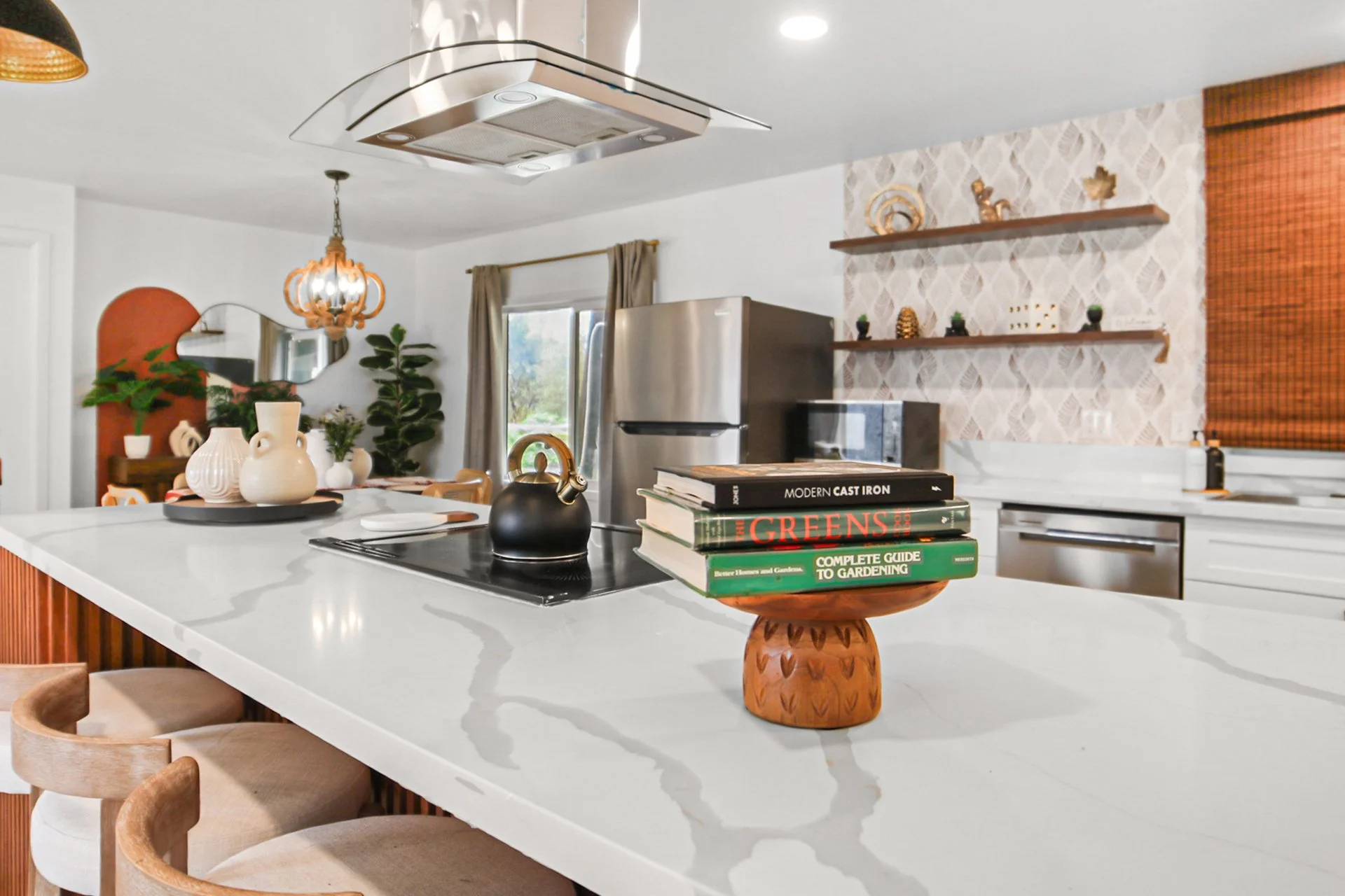 Modern kitchen with white island counter, decorative vases, and books on a wooden stand. Stainless steel refrigerator, patterned wallpaper, open shelving with decorative items, window with beige curtains, and various plants. Overhead lighting and a h