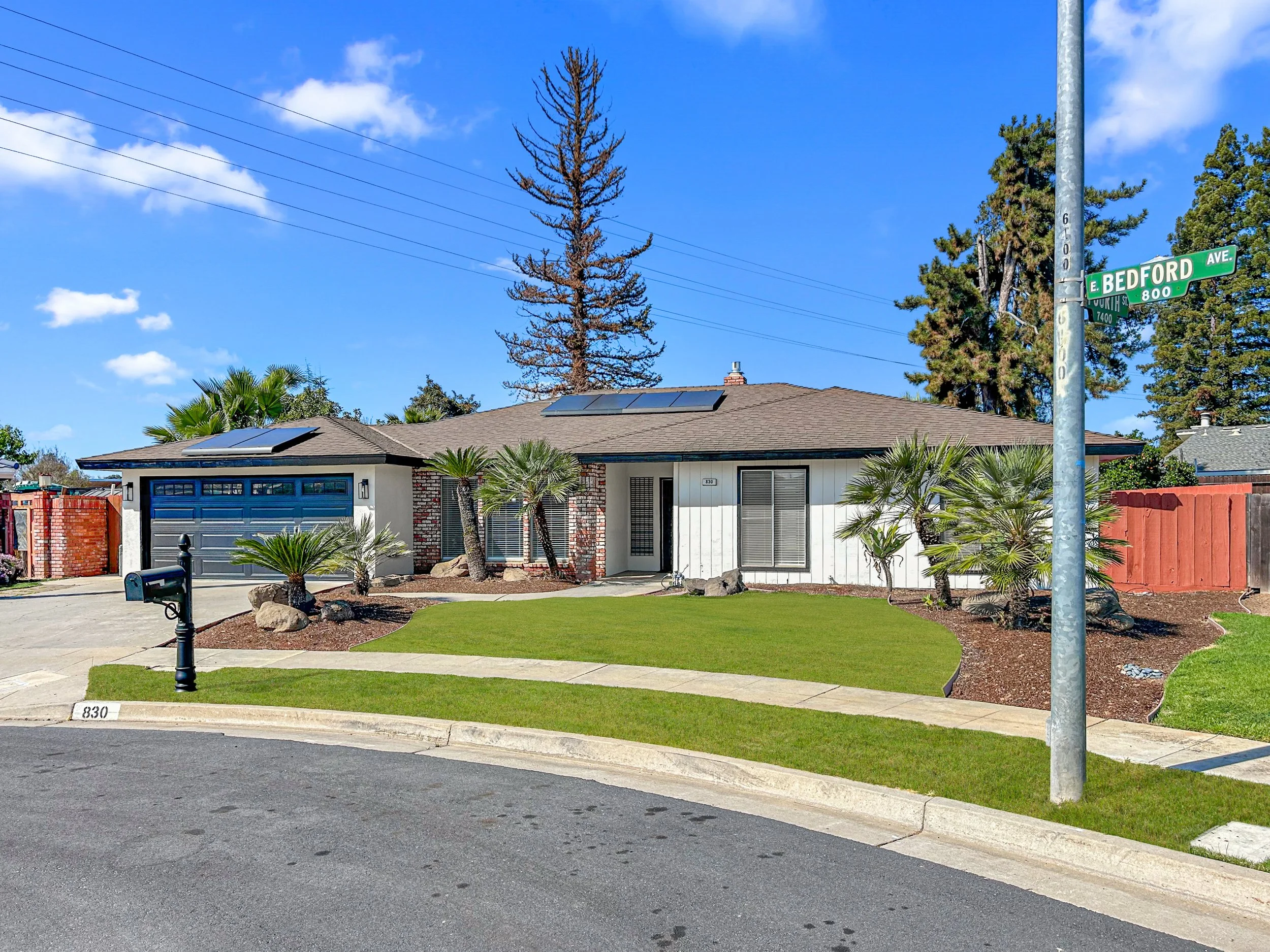 Single-story house at the corner of E Bedford Ave, with solar panels on the roof, a manicured lawn, palm trees, a mailbox, and a red wooden fence, under a blue sky with clouds.