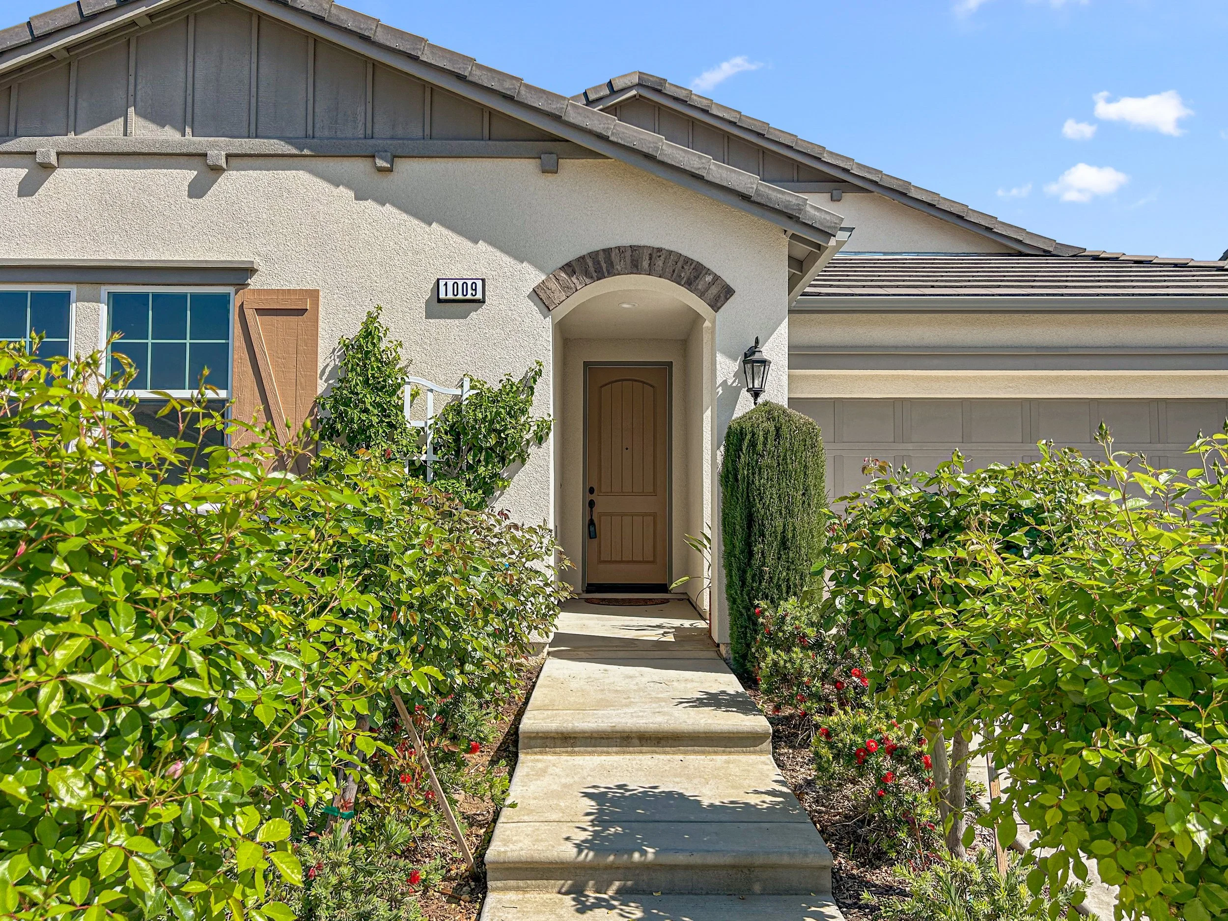 Front view of a house with a pathway through greenery leading to a wooden door. The house has a textured white facade, a brown window shutter, and a gray shingled roof. A black lantern light fixture is on the right of the door, and the house number 1