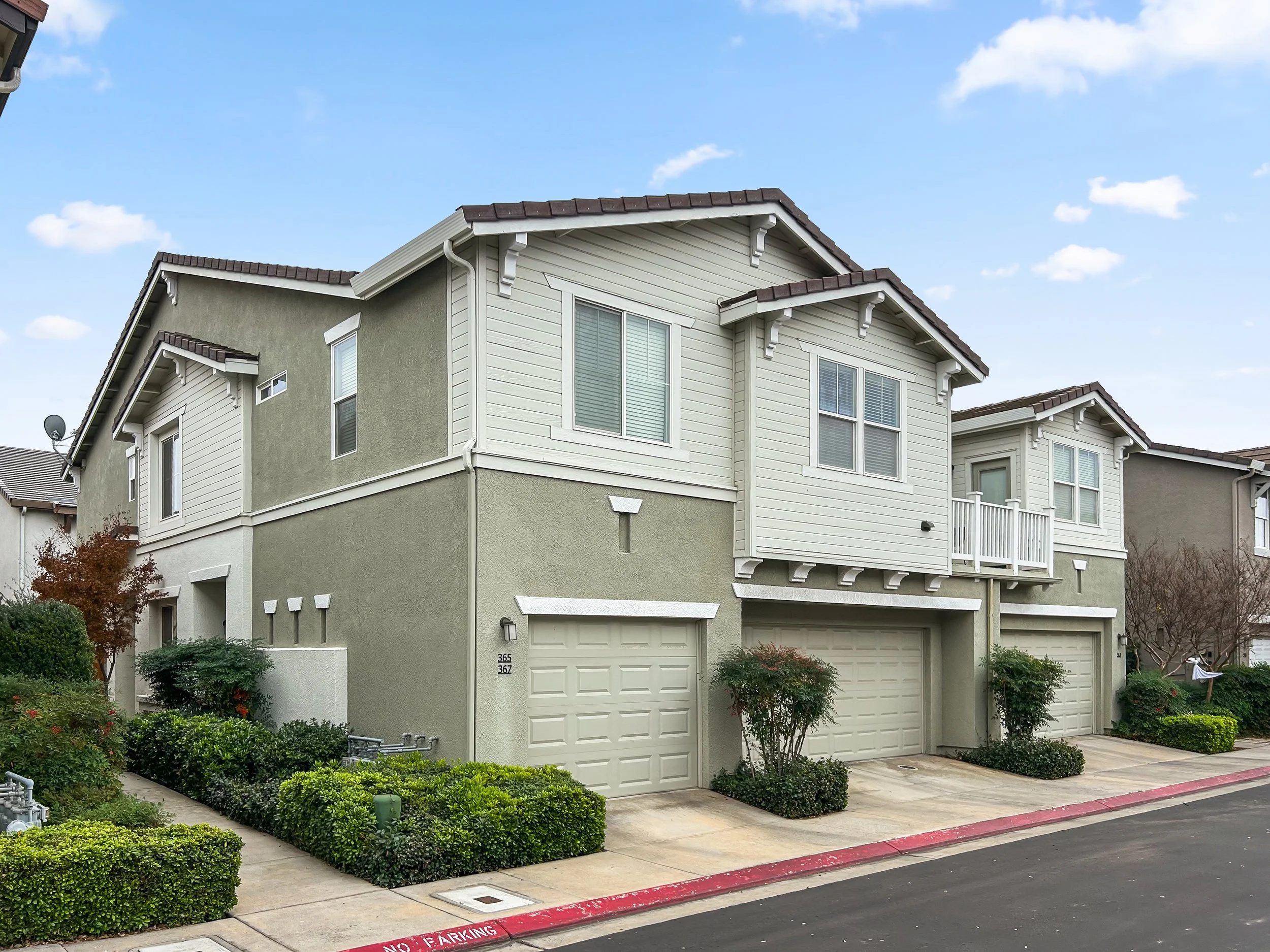 A multi-family residential building with three garage doors, landscaped bushes, trees, and a sidewalk in front.