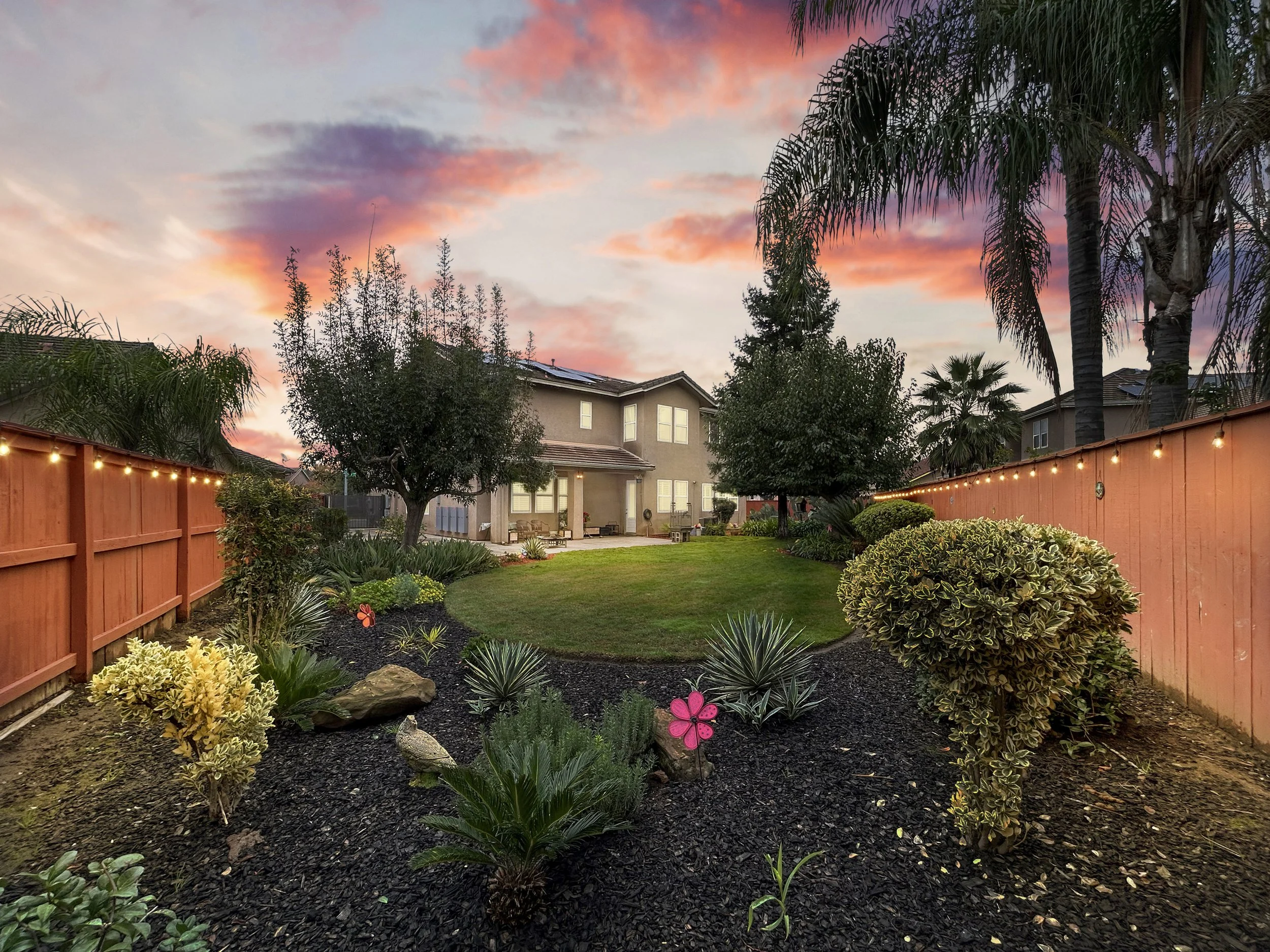 Backyard with a well-maintained lawn, colorful flowers, shrubs, trees, wooden fence with string lights, and a house in the background during sunset.