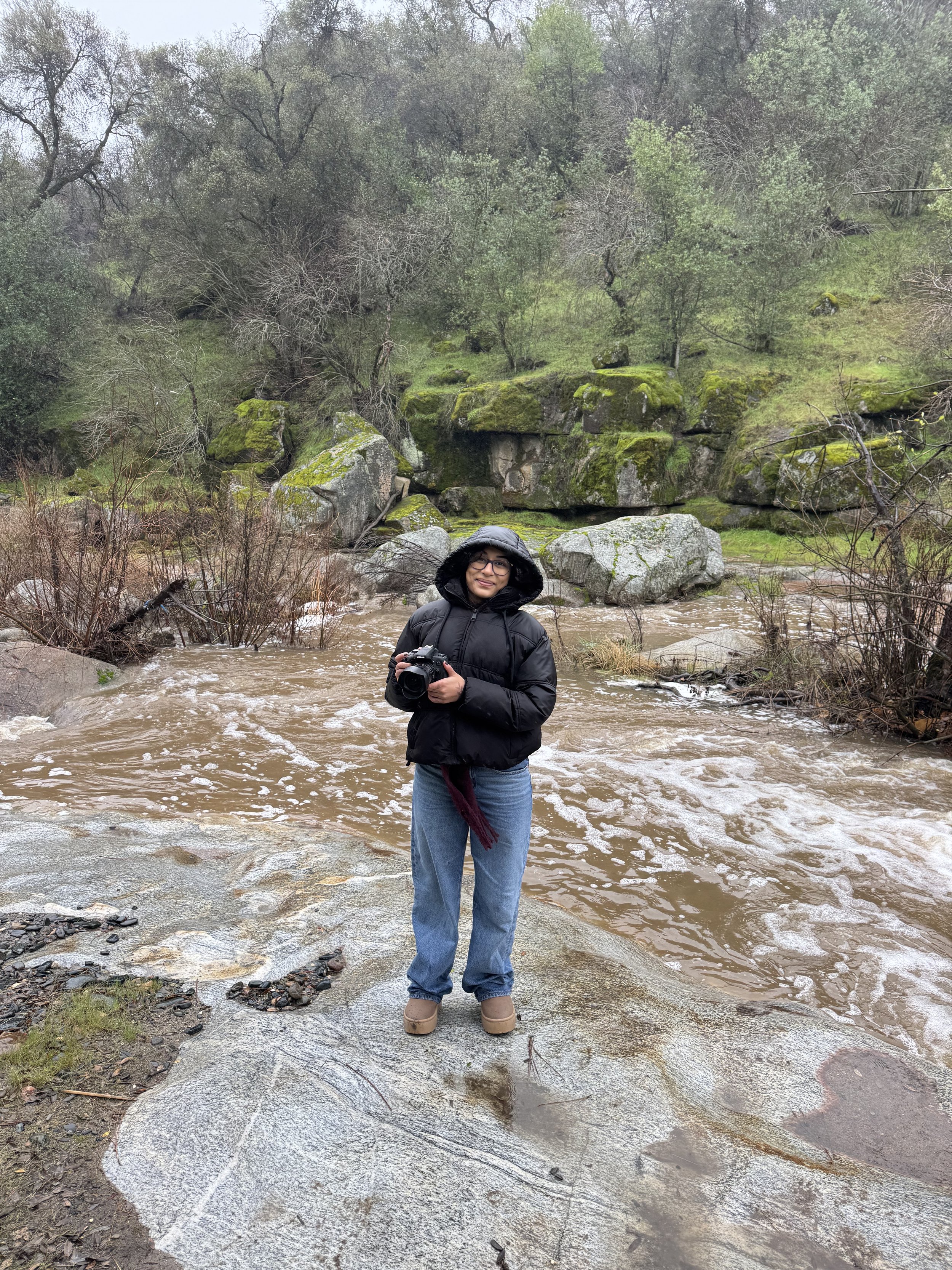 Person standing on wet rock holding a camera near a creek in a forest with moss-covered rocks and trees in the background.