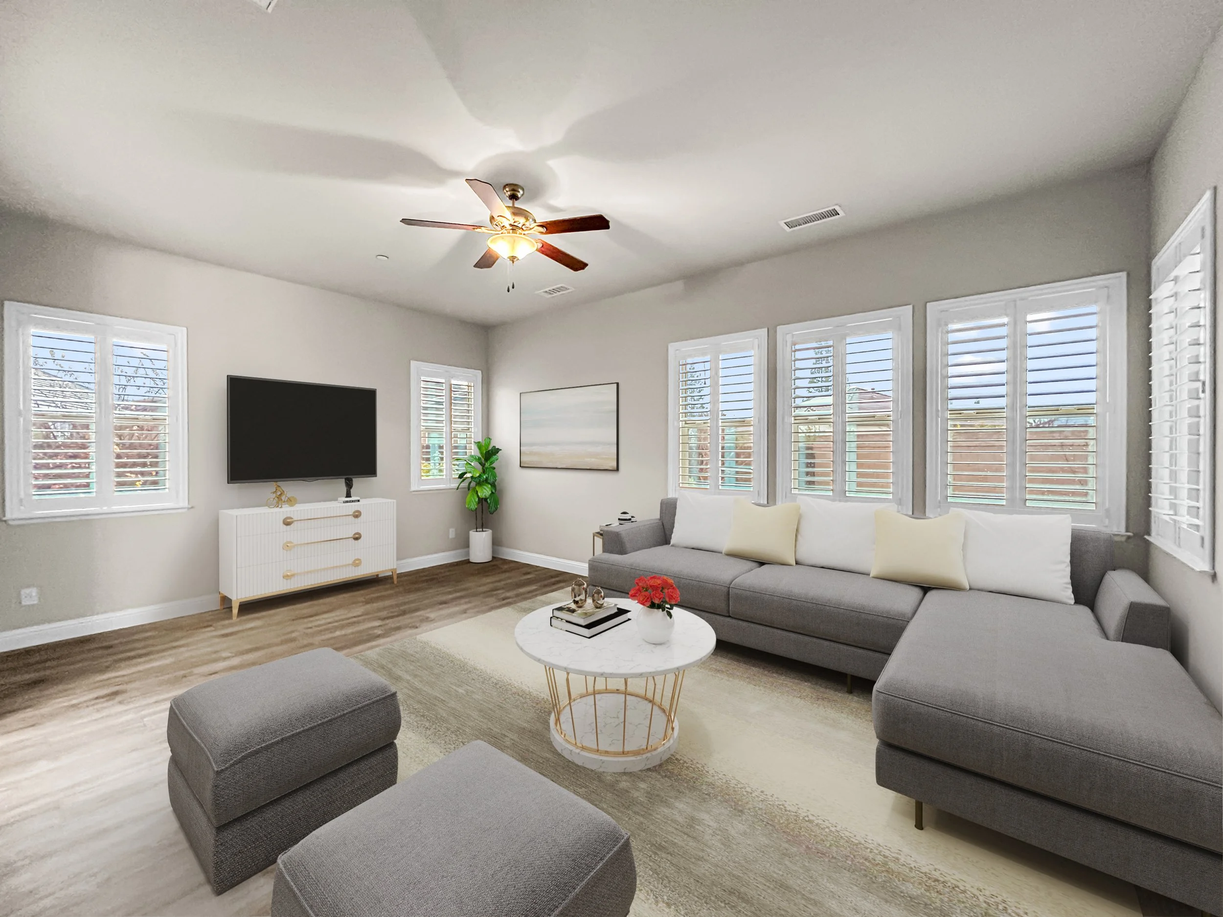 Modern living room with gray sectional sofa, white and yellow pillows, round white coffee table with red flowers, wall-mounted flat-screen TV, white dresser, potted plant, framed artwork, multiple windows with white shutters, and hardwood floors.
