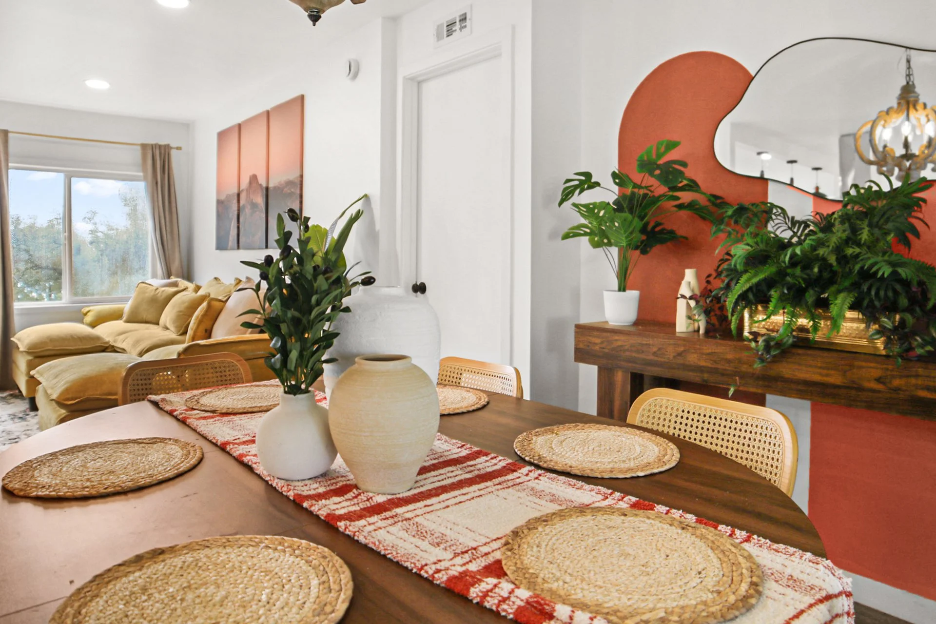 A dining area with a wooden table, woven placemats, and a red and white table runner, decorated with vases and greenery, blending into a living room with a yellow sofa, large window, and wall art.