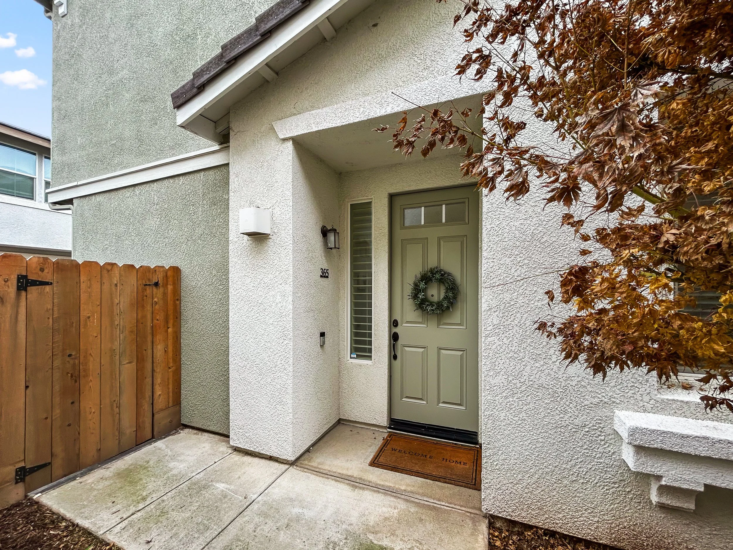 Front entrance of a house with a green door featuring a wreath, a welcome mat, and a small sidelight window. To the left, there is a wooden fence gate, a wall-mounted light, and the house number 365.
