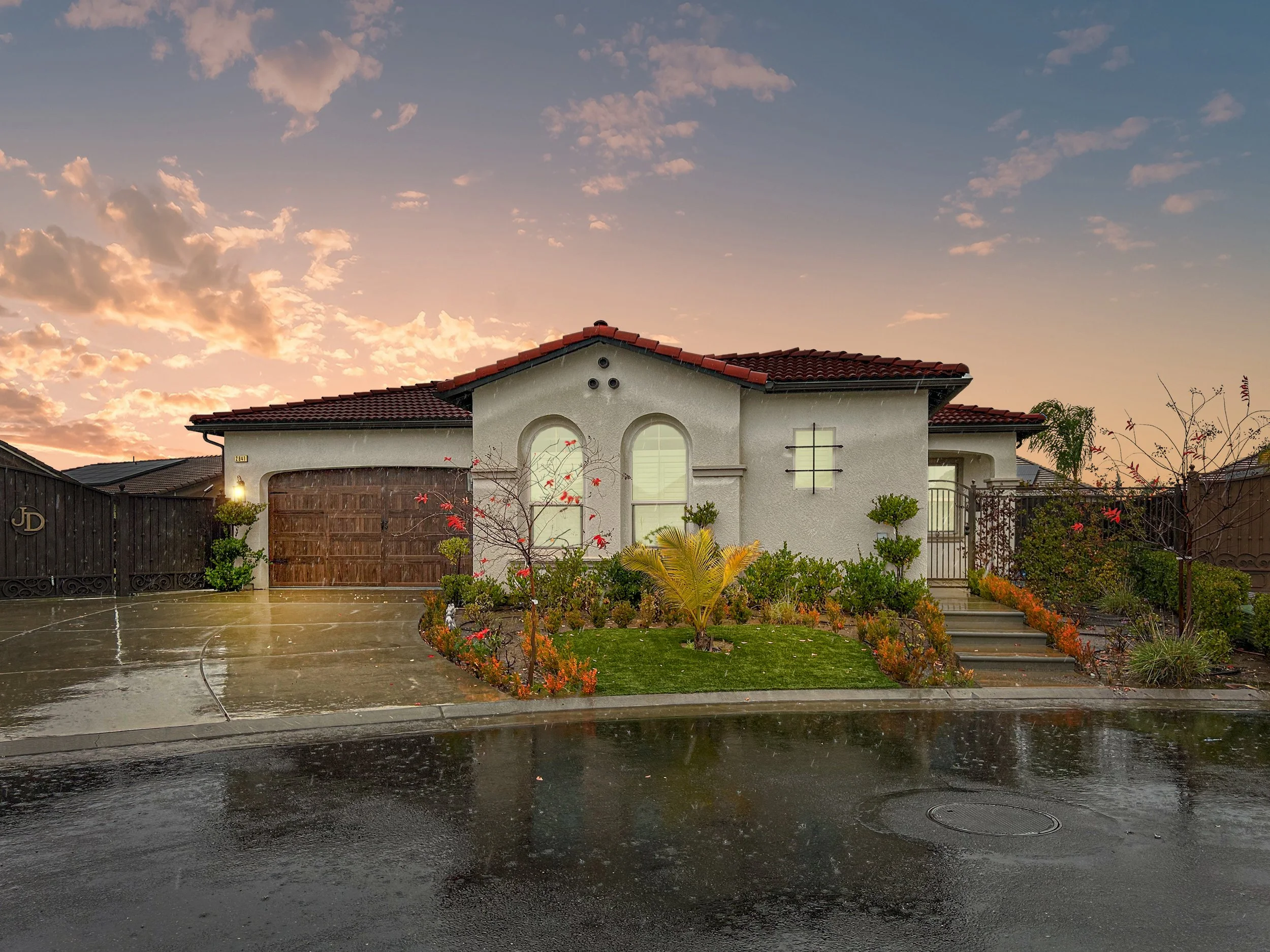 A modern single-story house with a red-tiled roof, surrounded by a garden and a wet driveway, at sunset with a partly cloudy sky.