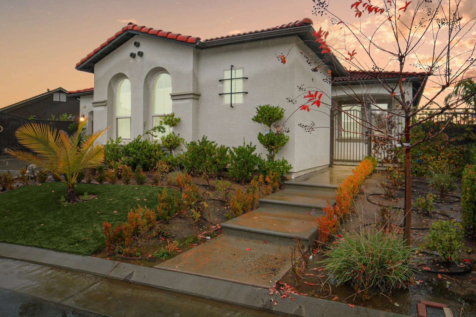 Front yard of a white house with a red-tiled roof, garden, and wet sidewalk at sunset.