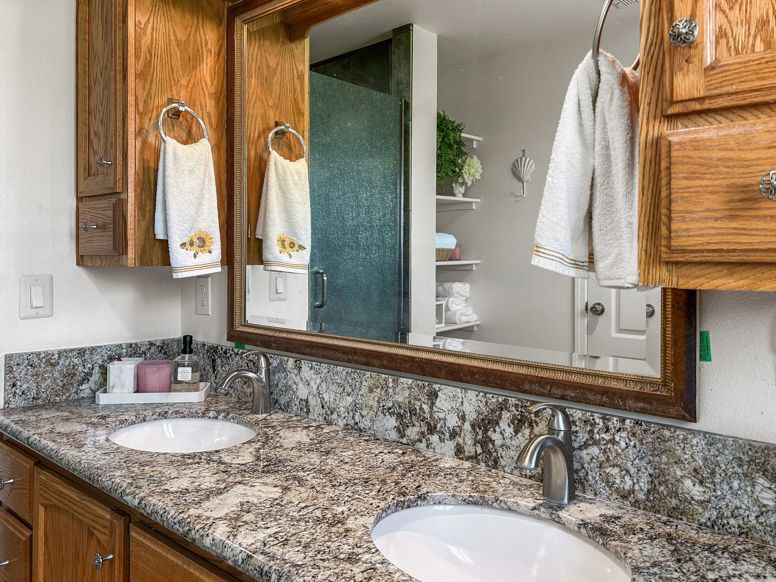 Bathroom with granite countertop, two undermount sinks, wooden cabinets, large mirror, towels hanging, shower in the background, shelves with towels and decorations.