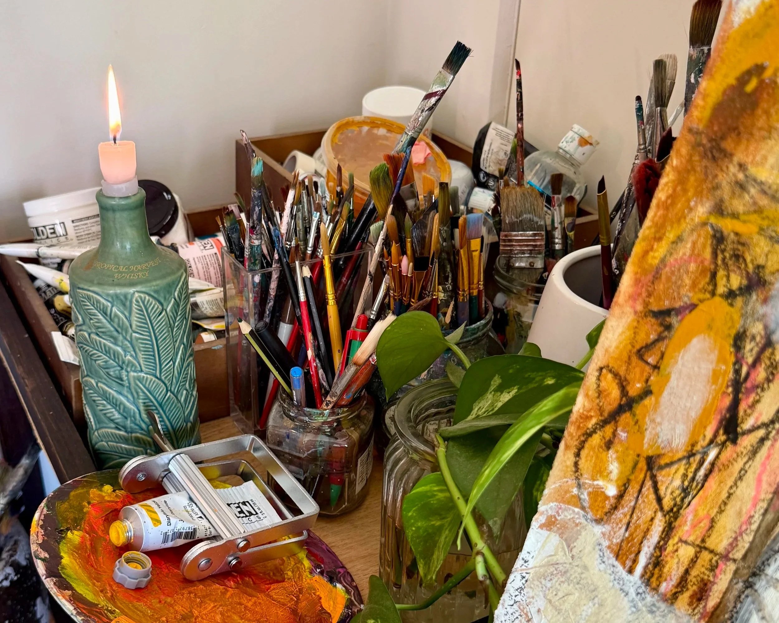 An artist's workspace with paintbrushes, paint tubes, a palette with orange and yellow paint, a candelabra with a pink candle, a potted plant, and abstract artwork.