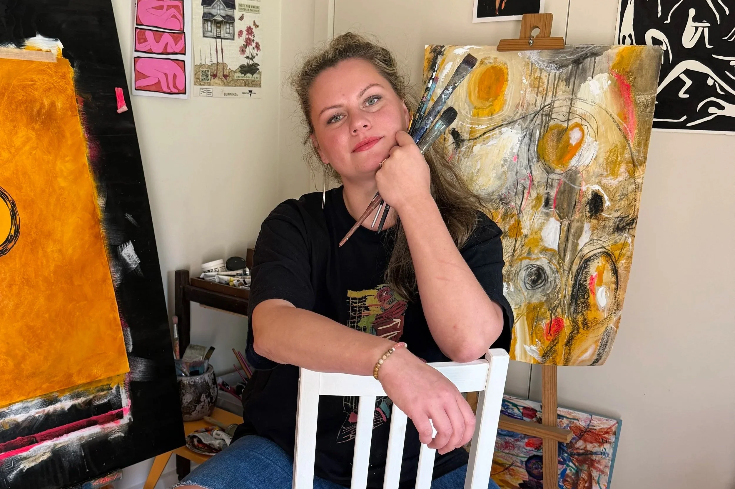 A woman with curly hair holding paintbrushes sitting in front of an abstract painting with yellow, black, and white colors, surrounded by additional art on the walls and workspace in an art studio.