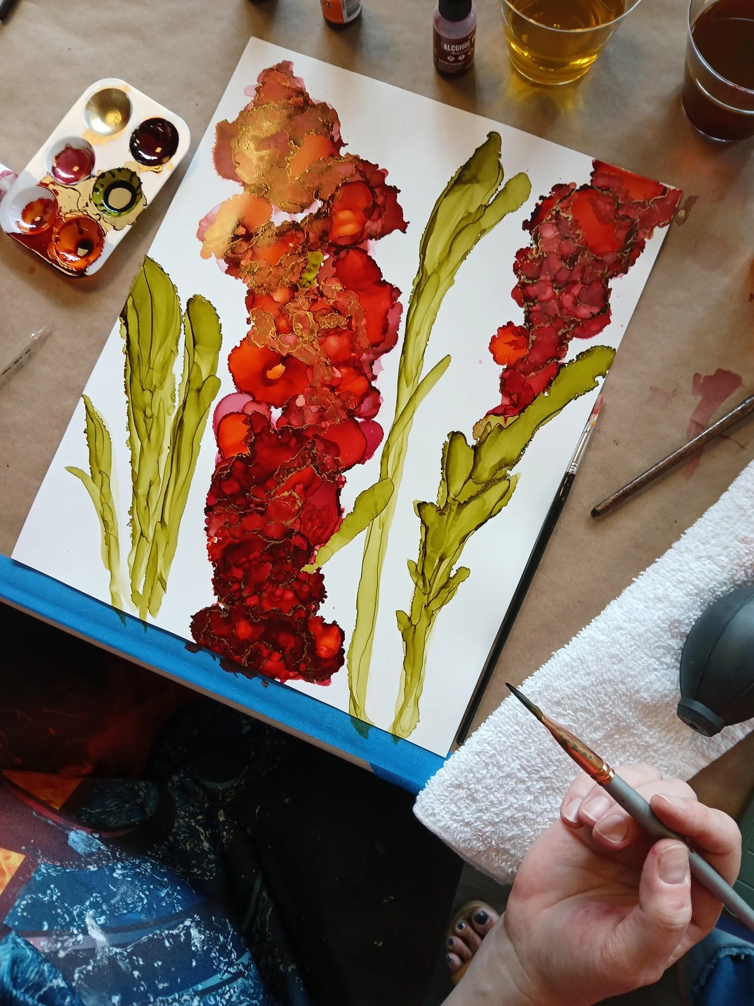 Alcohol ink painting of red flowers with green stems in progress, with ink palette and brushes