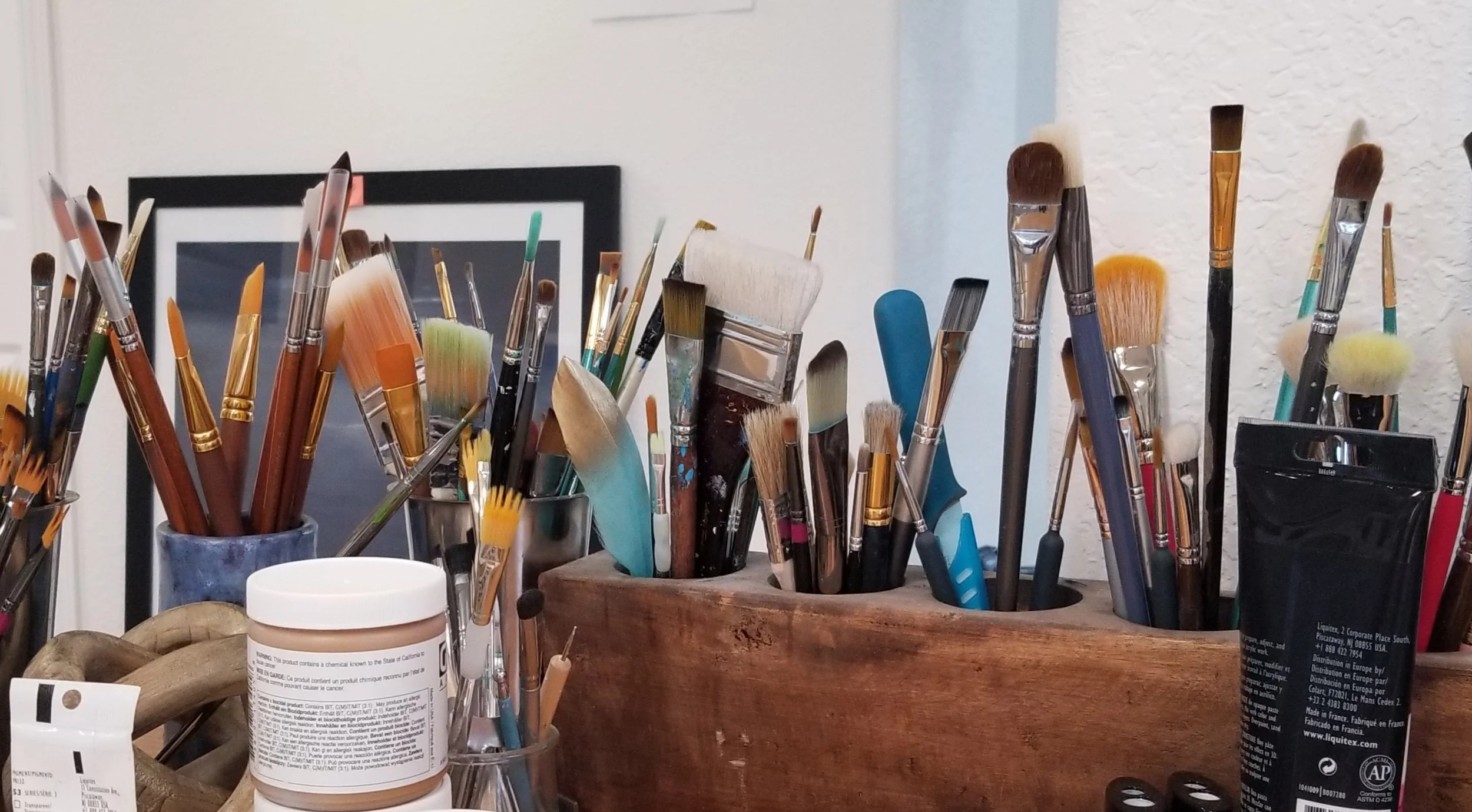 Paintbrushes of many sizes in a wooden holder and jars beside acrylic paint at T.Zinke Art Studio